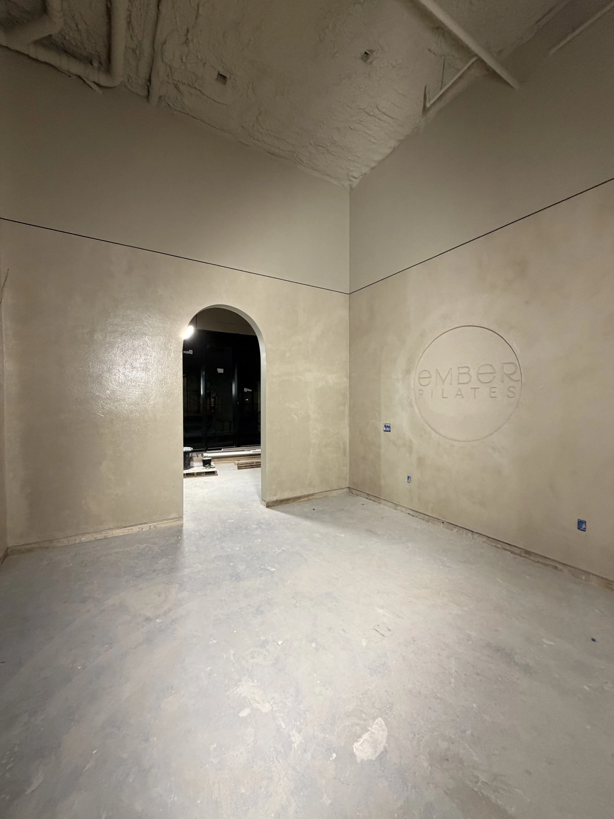 An indoor space with unfinished concrete flooring, beige walls, and a doorway leading to a room with construction materials. There is a circular logo on the wall that reads 'EMBER PILATES'.