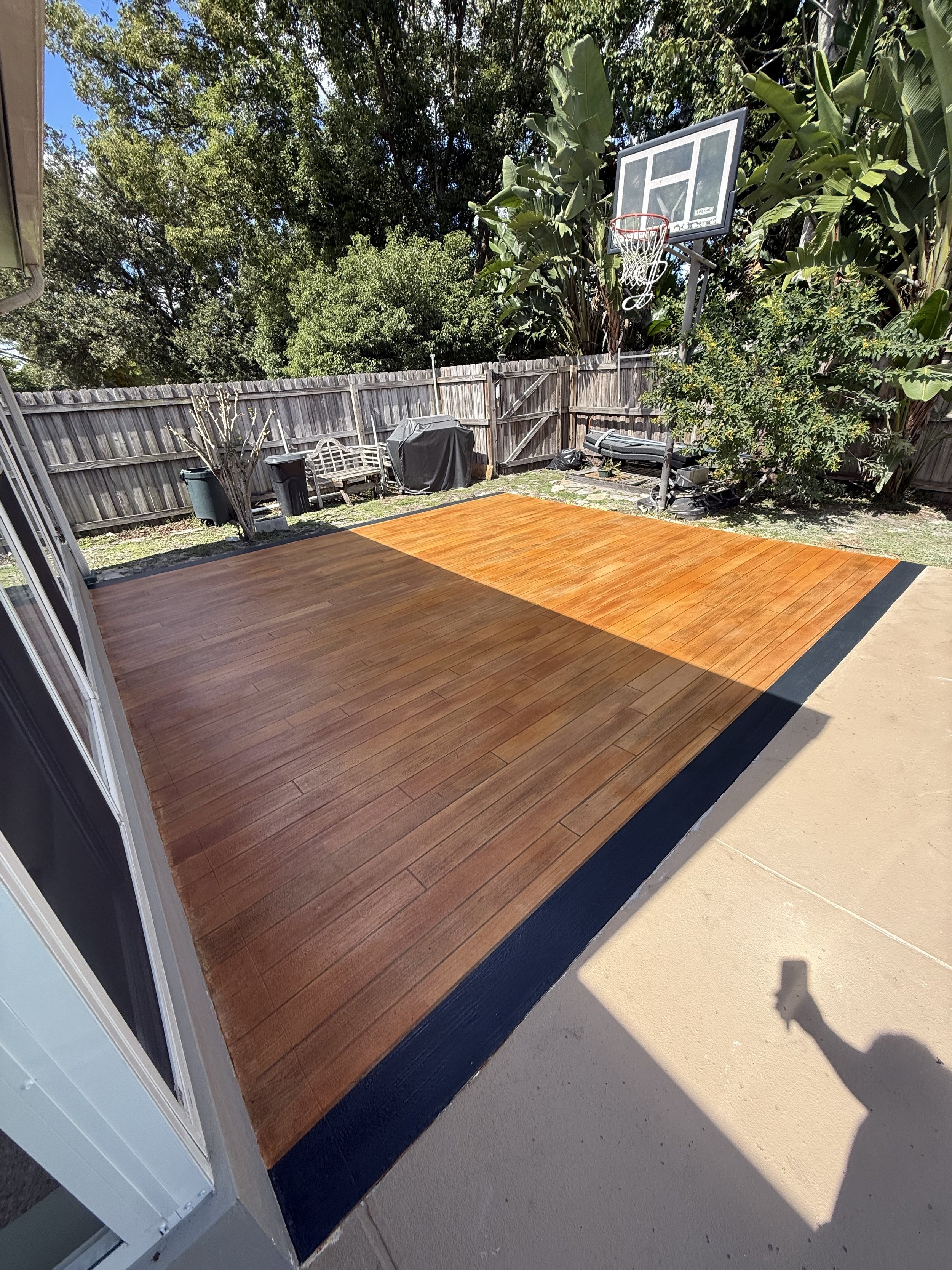 Backyard with a wooden deck, basketball hoop, outdoor furniture, grill, trash bins, and lush green trees and plants.
