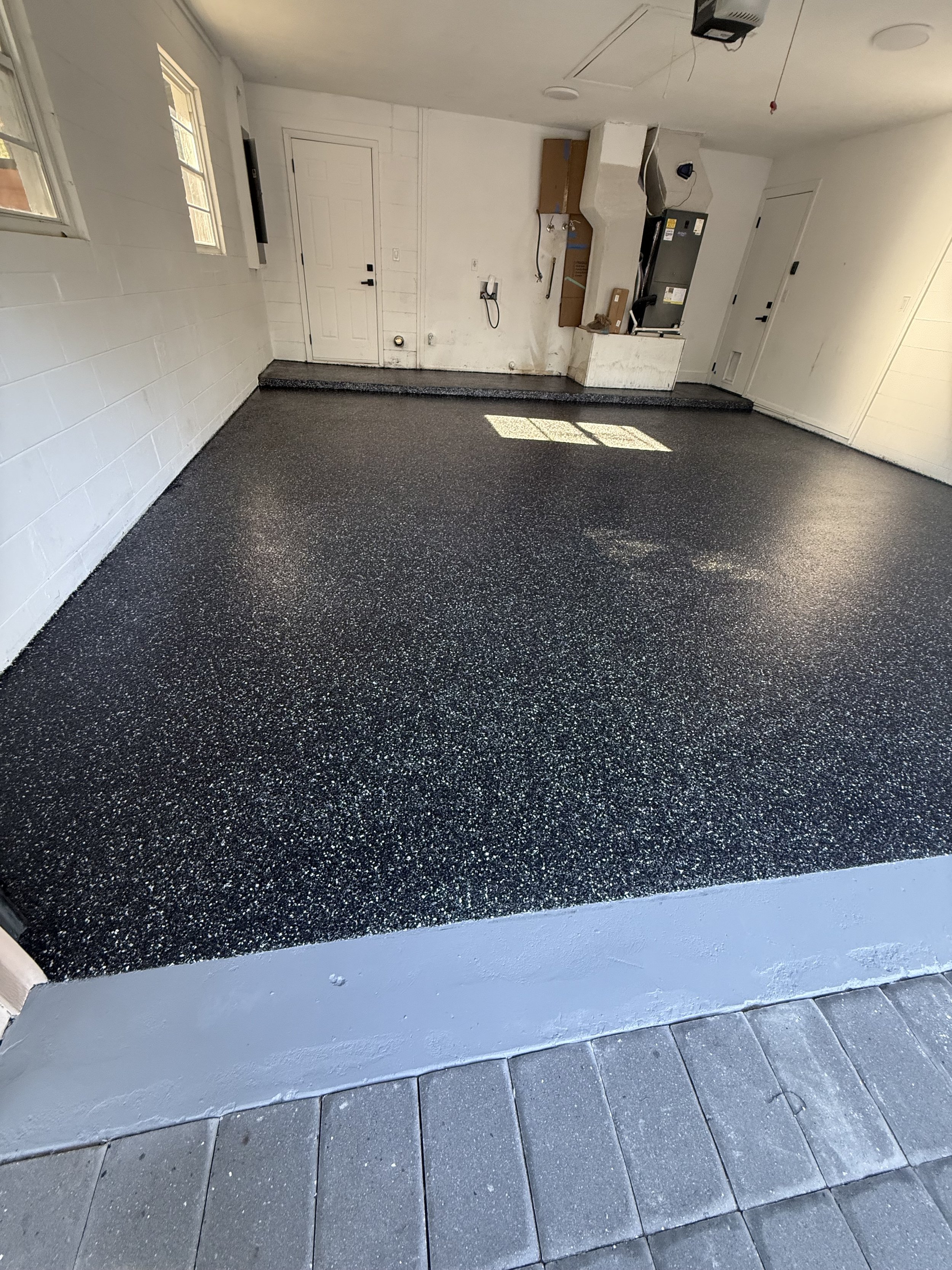 A room with a dark speckled epoxy floor, white walls, two small windows, and an interior door. A step leads to an area with a countertop and some electrical appliances and wiring, indicating construction or renovation work.