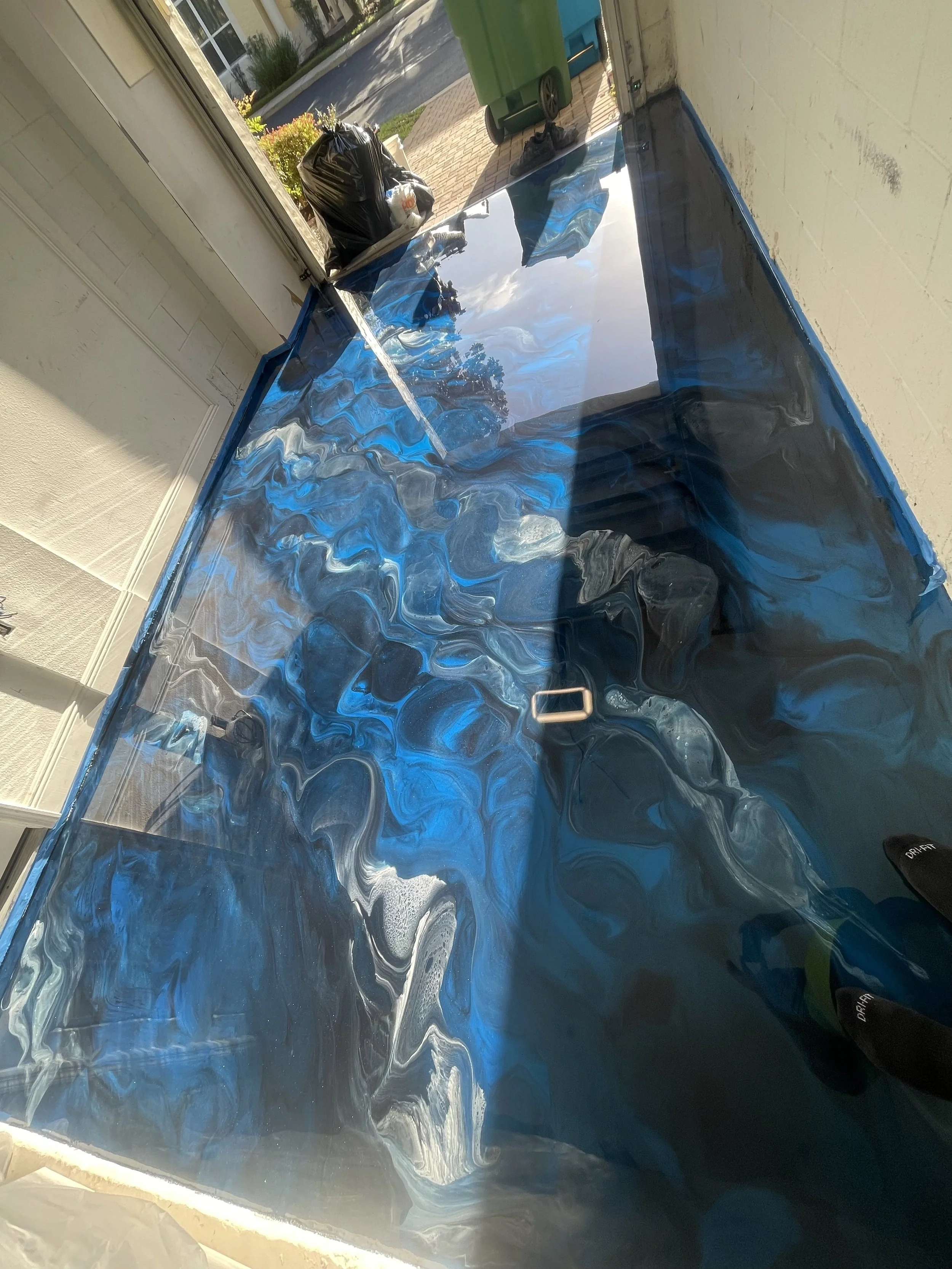 Residential garage with a partially painted blue marble-like patterned wall and some outdoor items including a green trash bin, black trash bag, shoes, and a plastic bottle, outside a driveway.