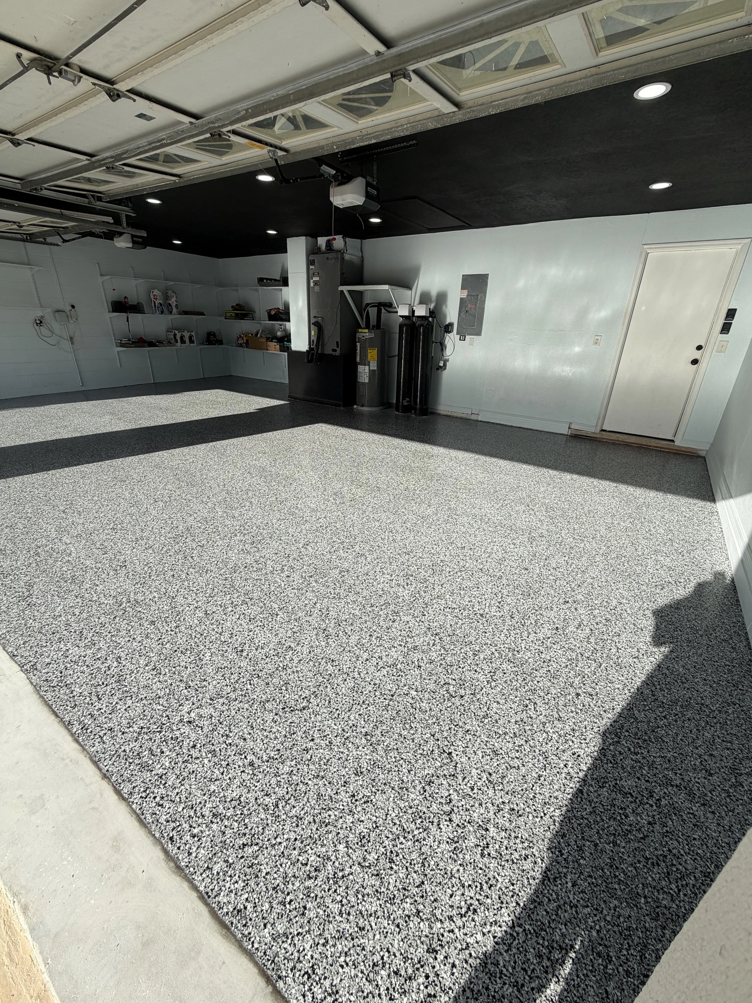 A garage with an open door, a section of the floor with black and white speckled epoxy coating, and tools and supplies on shelves along the wall.