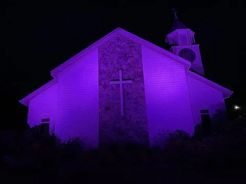 Church building illuminated with purple lights at night, featuring a cross on the front and a clock tower.