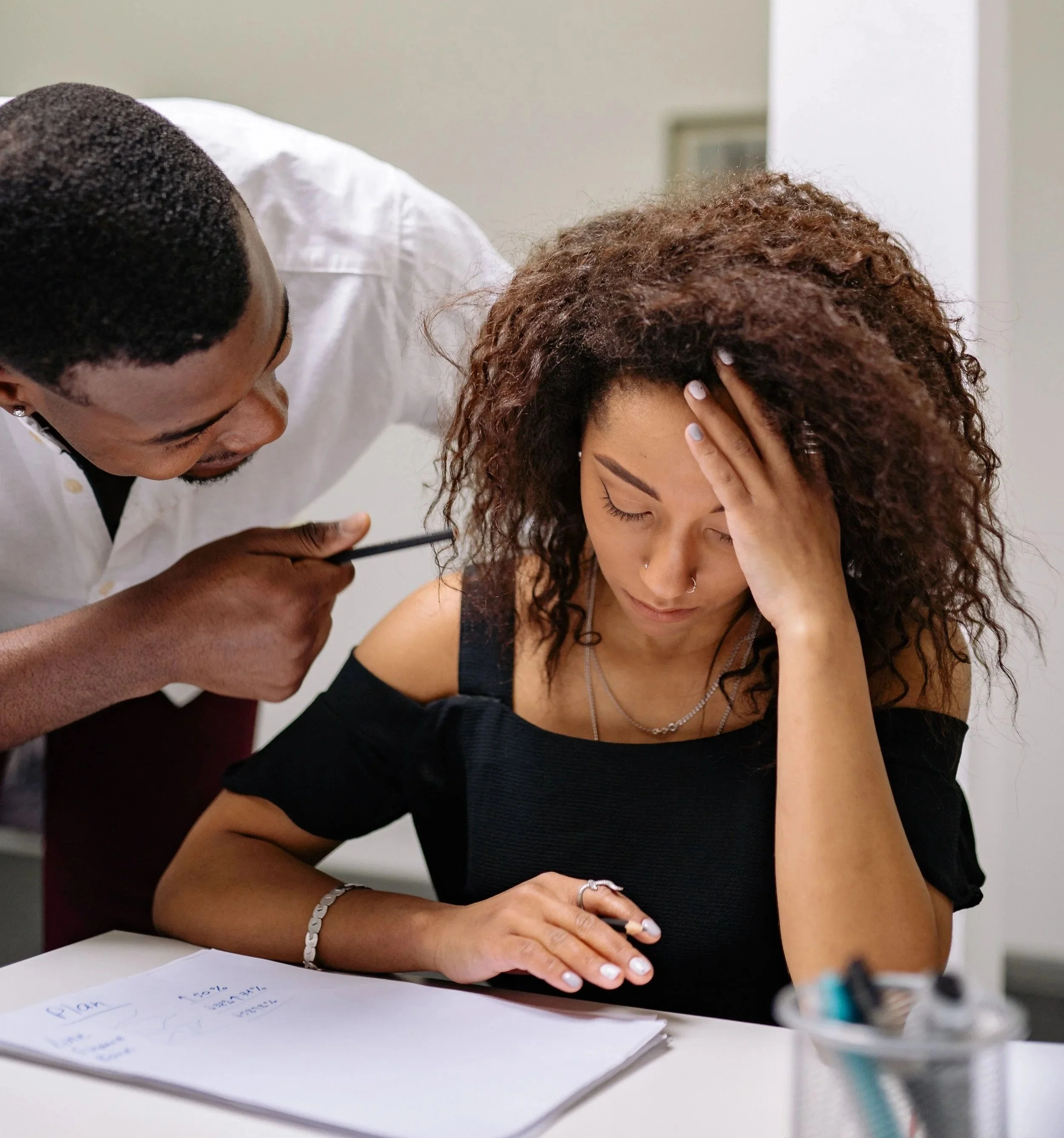 A man with short black hair leans over a woman with curly brown hair, who is resting her head on her hand while sitting at a table.