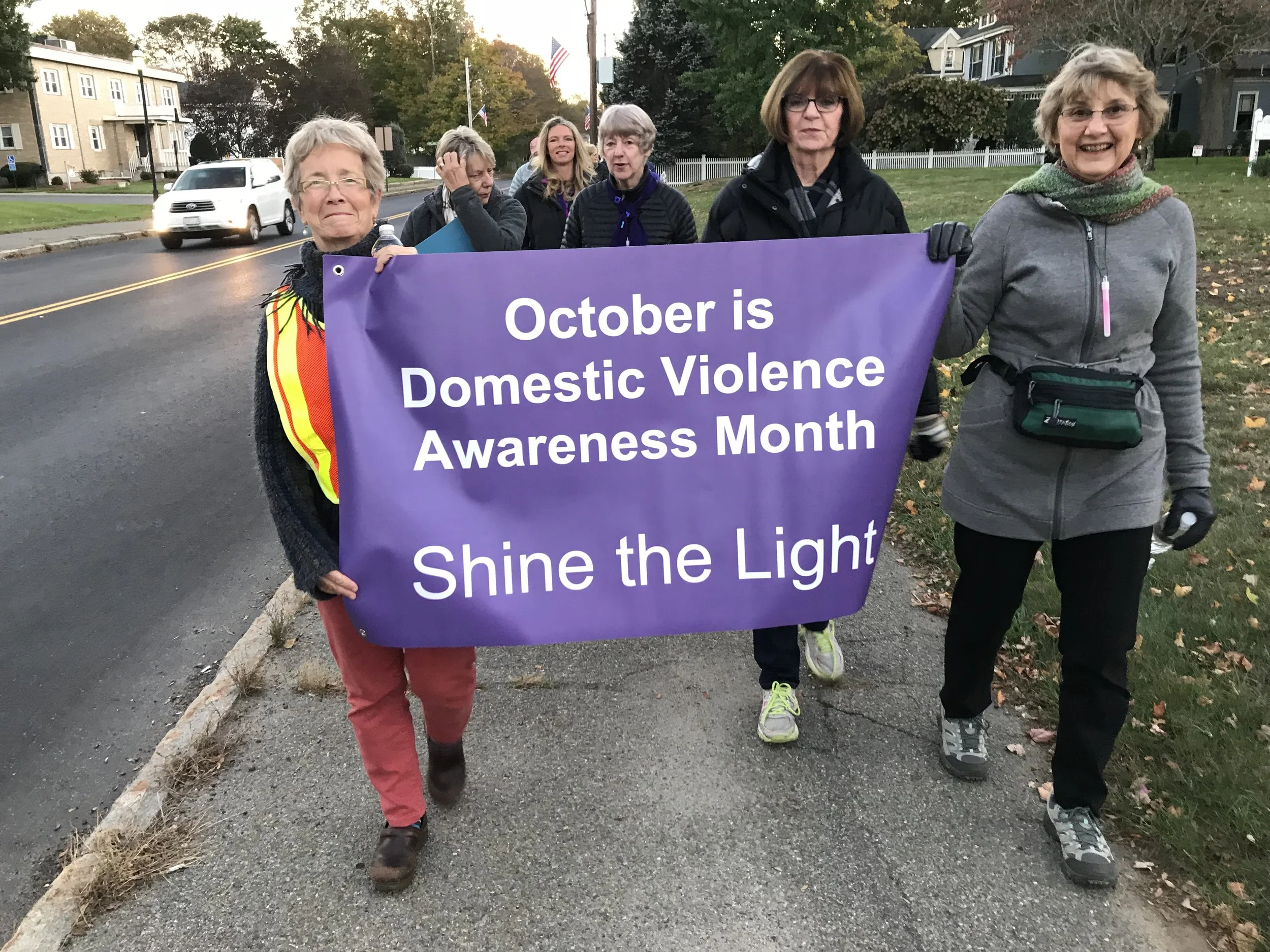 Group of women walking outdoors holding a purple banner that reads 'October is Domestic Violence Awareness Month' and 'Shine the Light'.