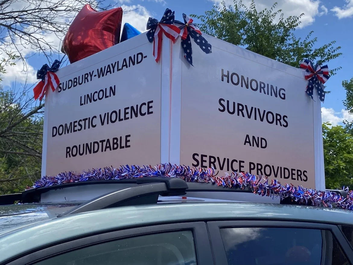 A float decorated with red, white, and blue ribbons and balloons, carrying a sign that reads, 'Subury-Wayland Lincoln Domestic Violence Roundtable, Honoring Survivors and Service Providers,' during a parade or community event.