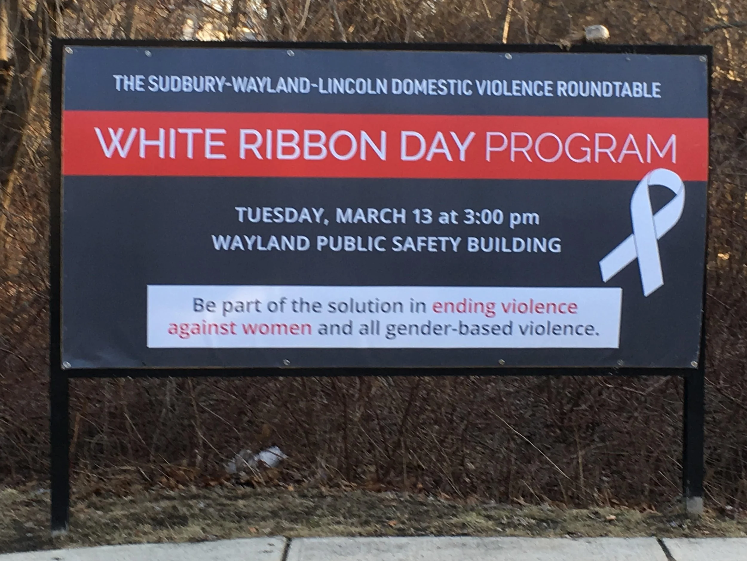 A signboard for the White Ribbon Day Program organized by the Sudbury-Wayland-Lincoln Domestic Violence Roundtable with a white ribbon symbol on the bottom right.
