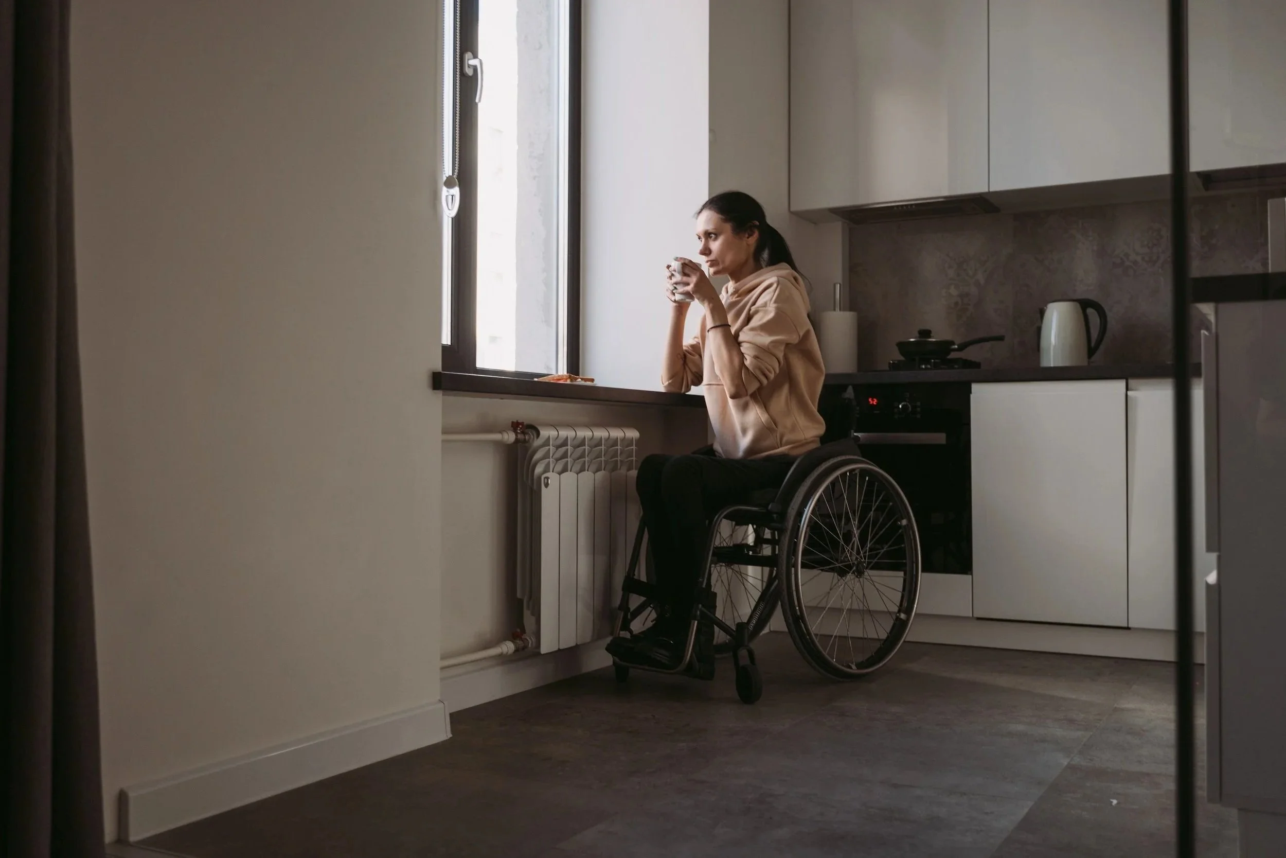 A woman in a beige hoodie sitting in a wheelchair, holding a mug, in a modern kitchen near a window.