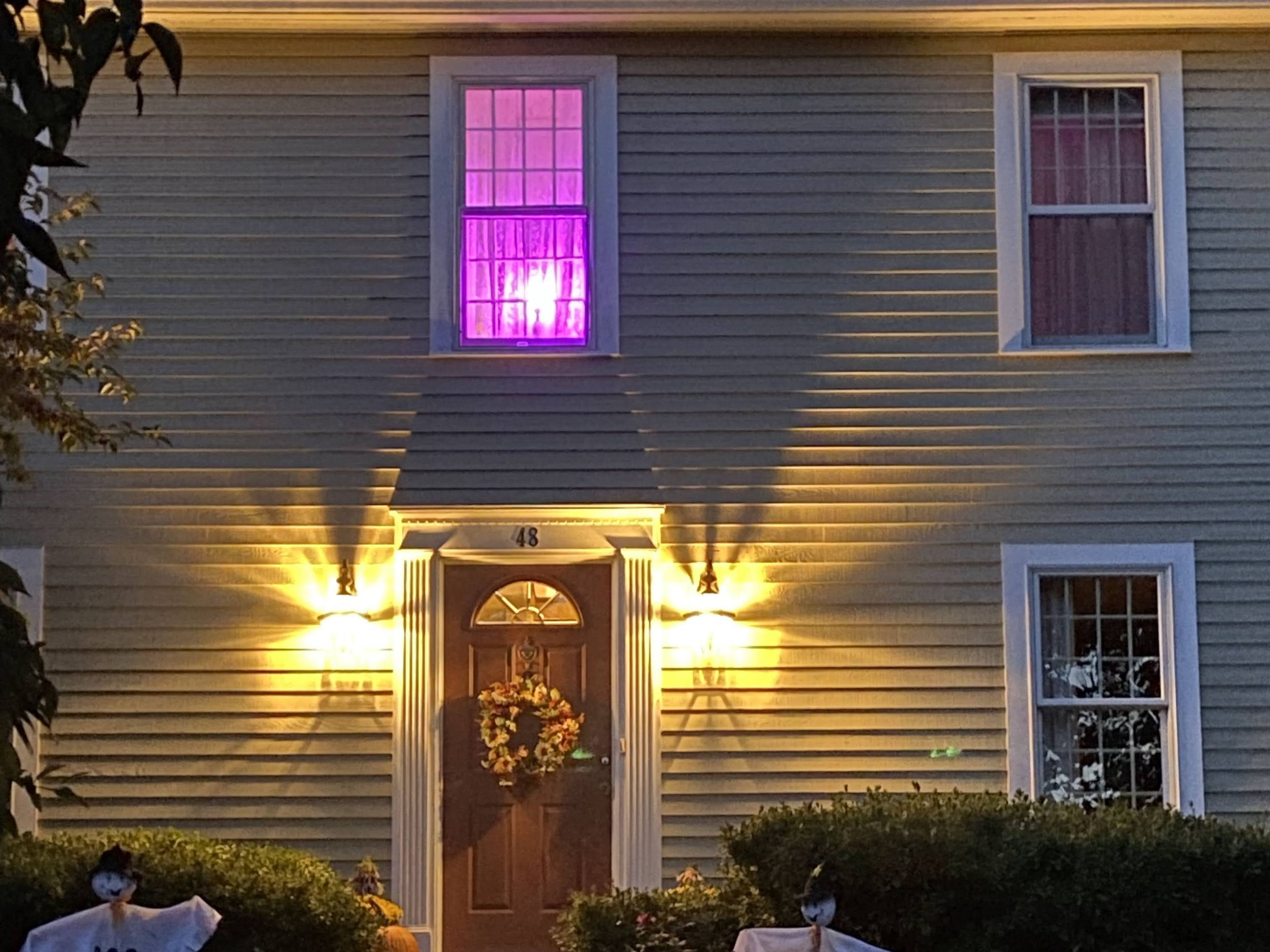 A house decorated for Halloween with purple-lit windows, a wreath on the front door, and Halloween decorations near the bushes.