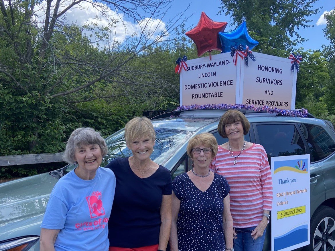 Four women standing in front of a decorated vehicle participating in a domestic violence awareness event, with signs and balloons supporting survivors and service providers.