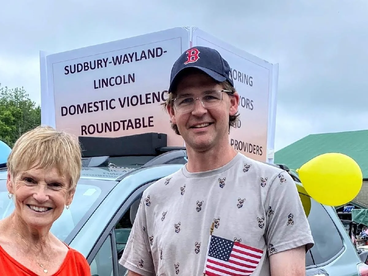 A woman and a man standing outdoors in front of a car with a sign that reads 'Sudbury-Wayland-Lincoln Domestic Violence Roundtable'.