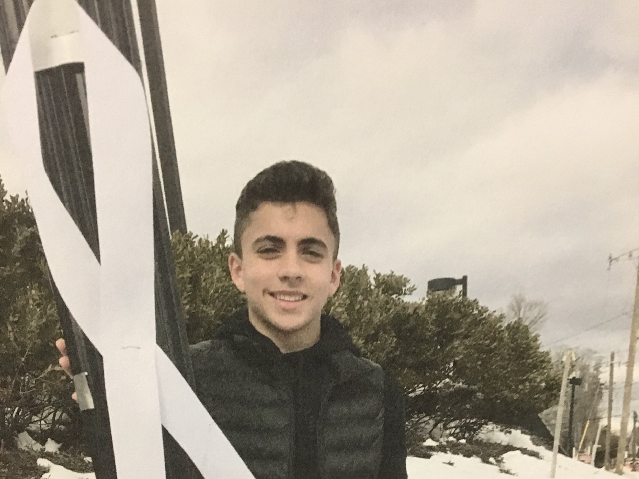 A young man smiling outdoors, raising his arms with a white ribbon beside him.