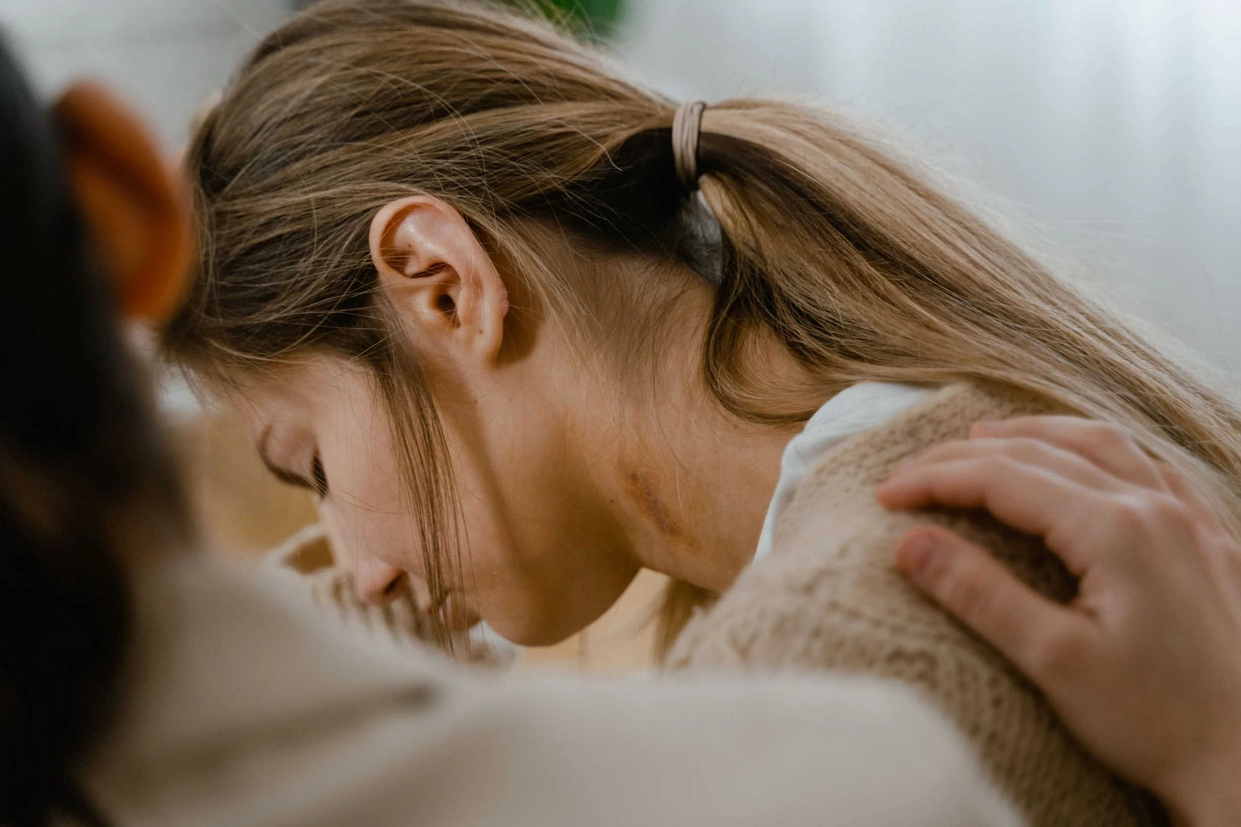 A woman with light brown hair tied in a ponytail and bent forward, with her head resting on her arm and hands on her shoulder.