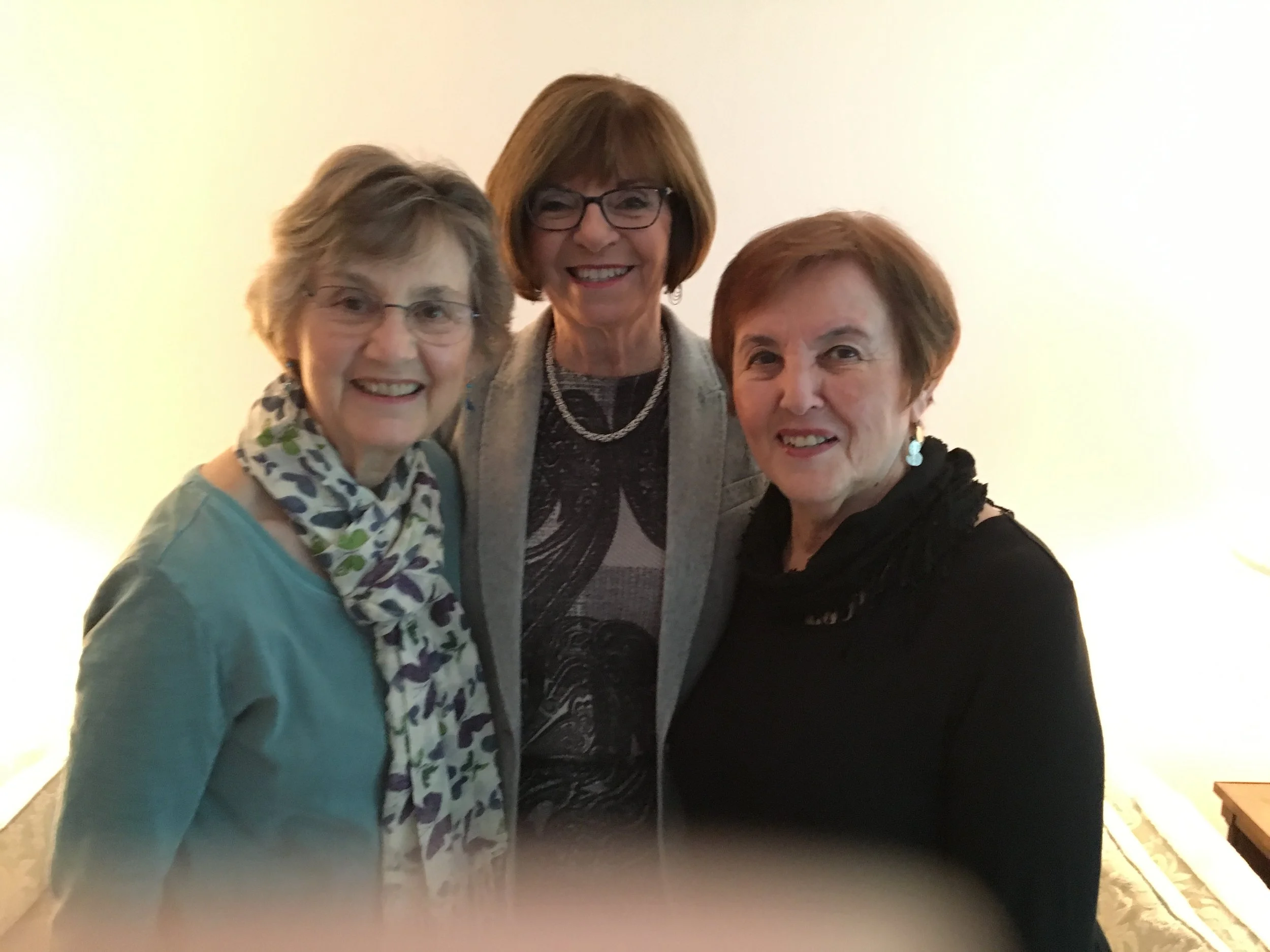 A group of three women smiling, standing close together, against a plain, light-colored background. They are wearing glasses and casual clothing.