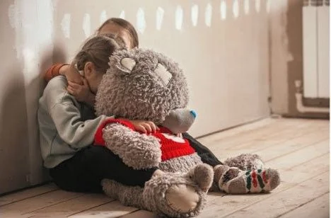 Two children hugging a large teddy bear sitting on the floor in a hallway.