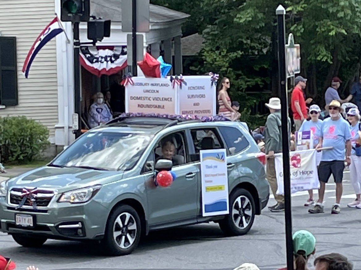 A parade scene with a gray SUV decorated with red, white, and blue balloons and ribbons, driving past a group of people holding banners.