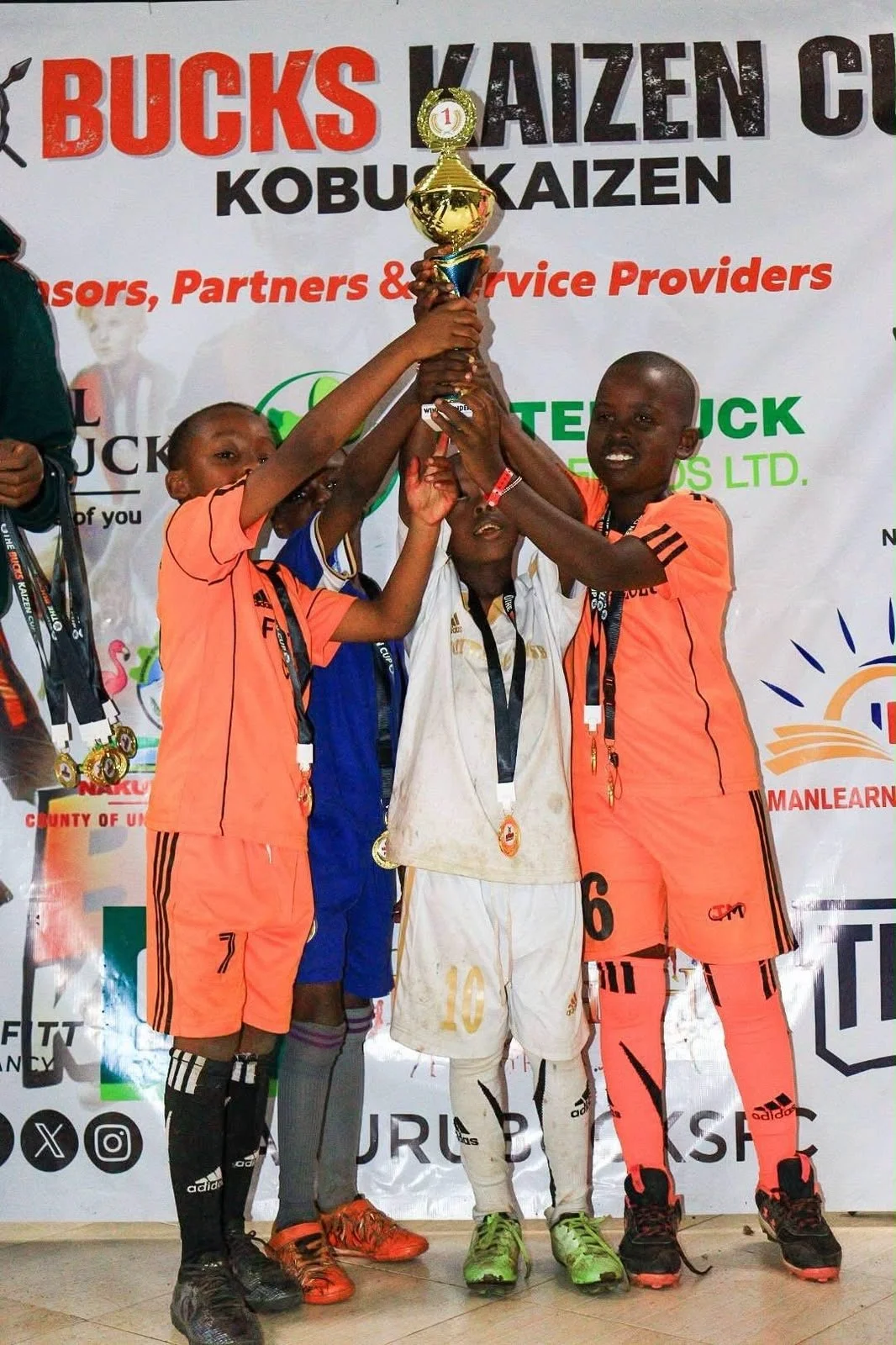 Young boys in sports uniforms celebrate on a podium, holding a trophy together during an award ceremony.