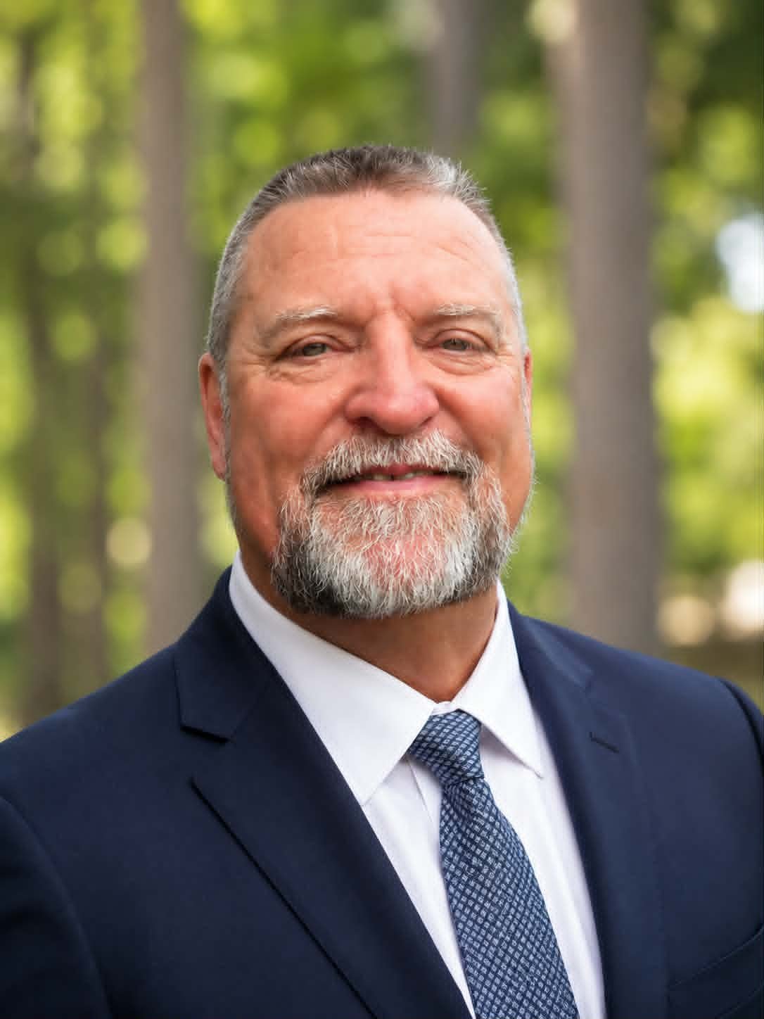 A middle-aged man with gray hair and a beard, dressed in a dark suit, white shirt, and patterned tie, standing outdoors with a blurry green forest background.
