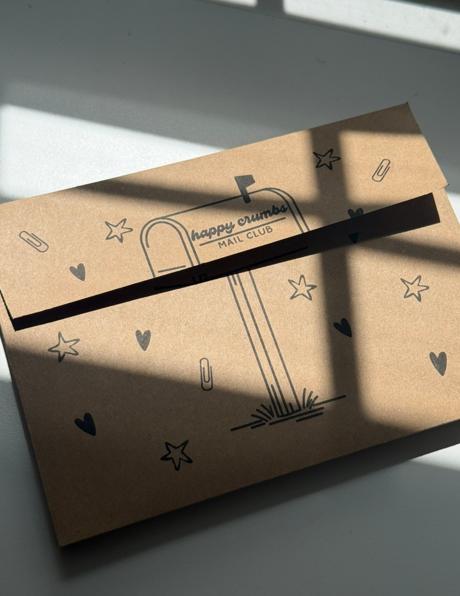 Brown Envelope with black doodles of stars, hearts, a mailbox, and paperclip symbols, and the words 'happy crumb MAIL CLUB' printed on it, placed on a white surface with shadows.