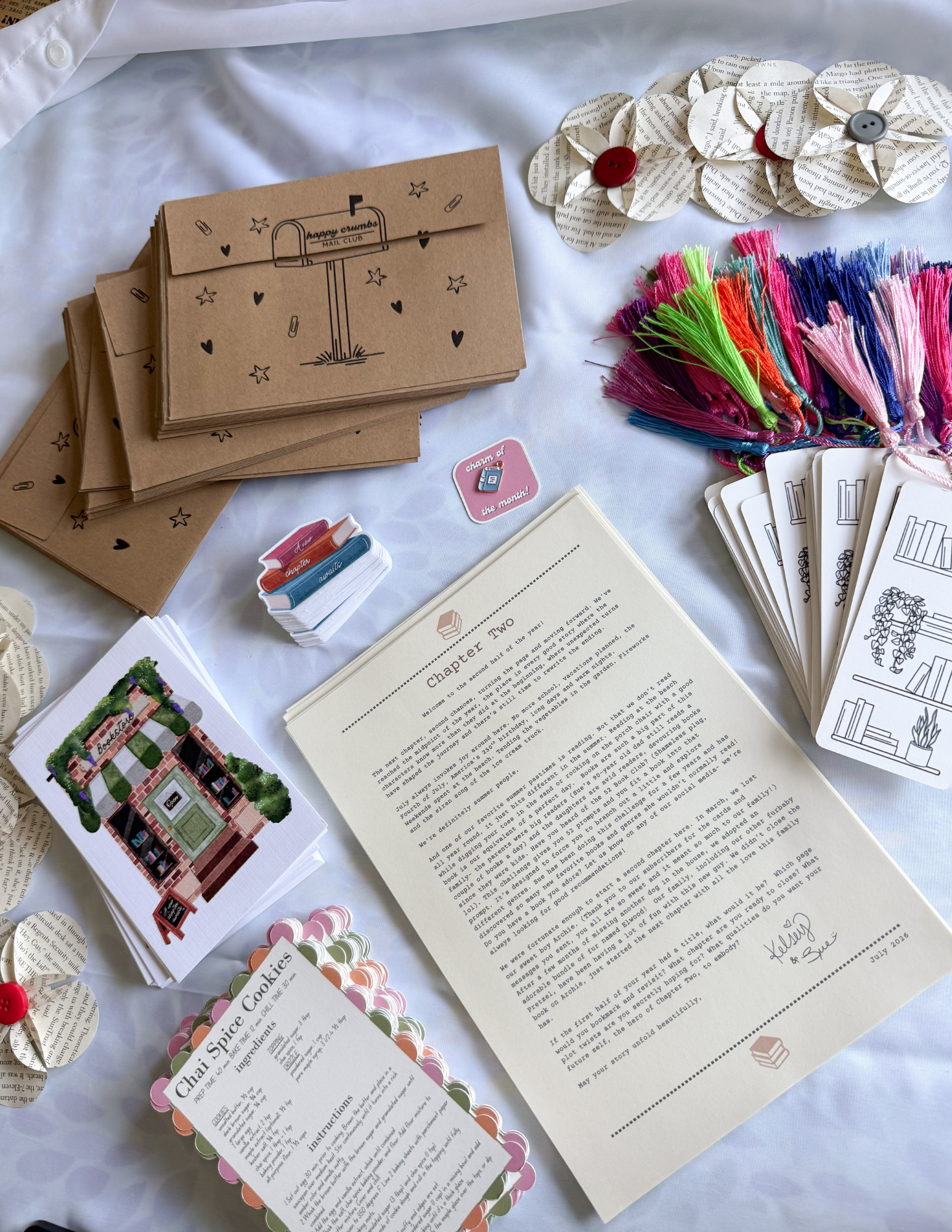 Display of paper crafts including paper flowers with button centers, colorful embroidery threads, illustrated postcards, printed book pages, and decorative stickers on a white fabric surface.