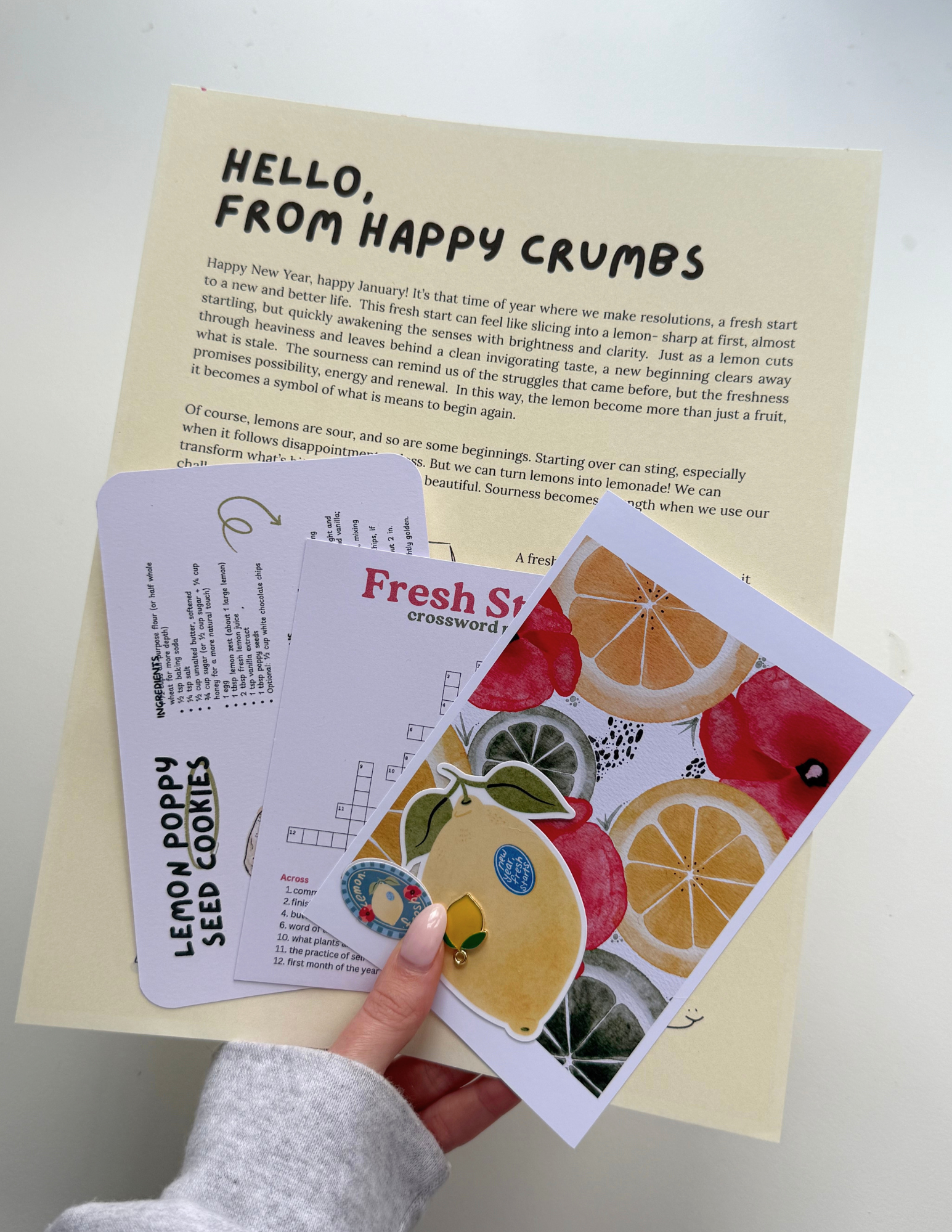 A person holding a colorful fruit-themed sticker and a crossword puzzle card, with a sheet of paper with text in the background.