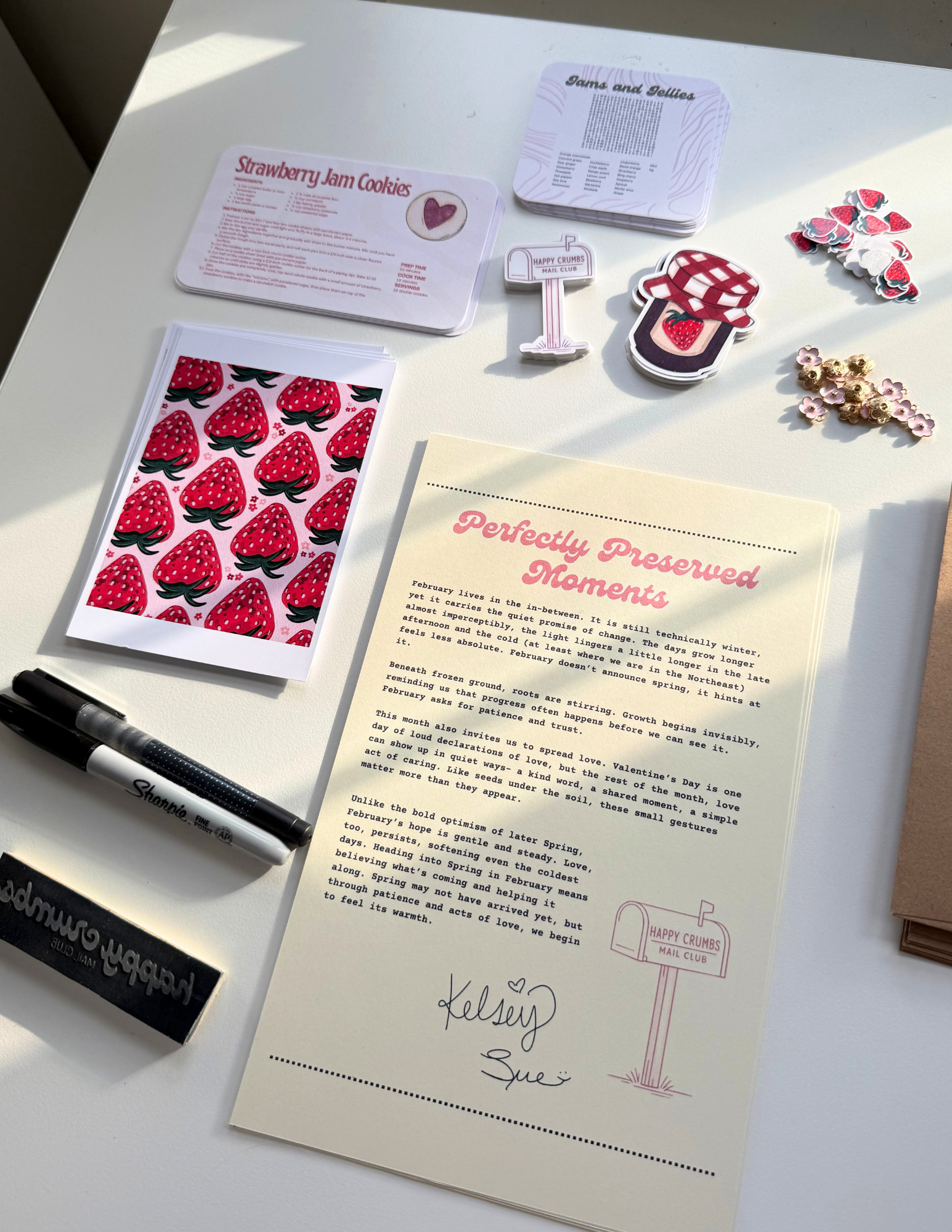 The image shows a white desk with various items spread out, including a stack of recipe cards for strawberry jam cookies, a notebook titled 'Perfectly Preserved Moments,' a black Sharpie marker, decorative stickers of strawberries and flowers, and a paper sign for 'Happy Crumbs Mail Club.' There is a handwritten note with the names 'Kelsey & Sue' and a heart symbol.