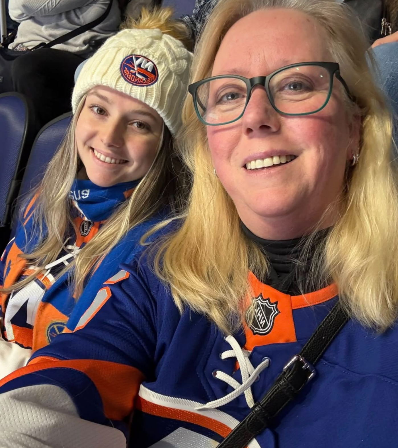 Two women, one younger and one older, wearing hockey jerseys and sitting on stadium seats at an arena. The younger woman is wearing a yellow knit hat with a logo, and the older woman has glasses. Both are smiling.