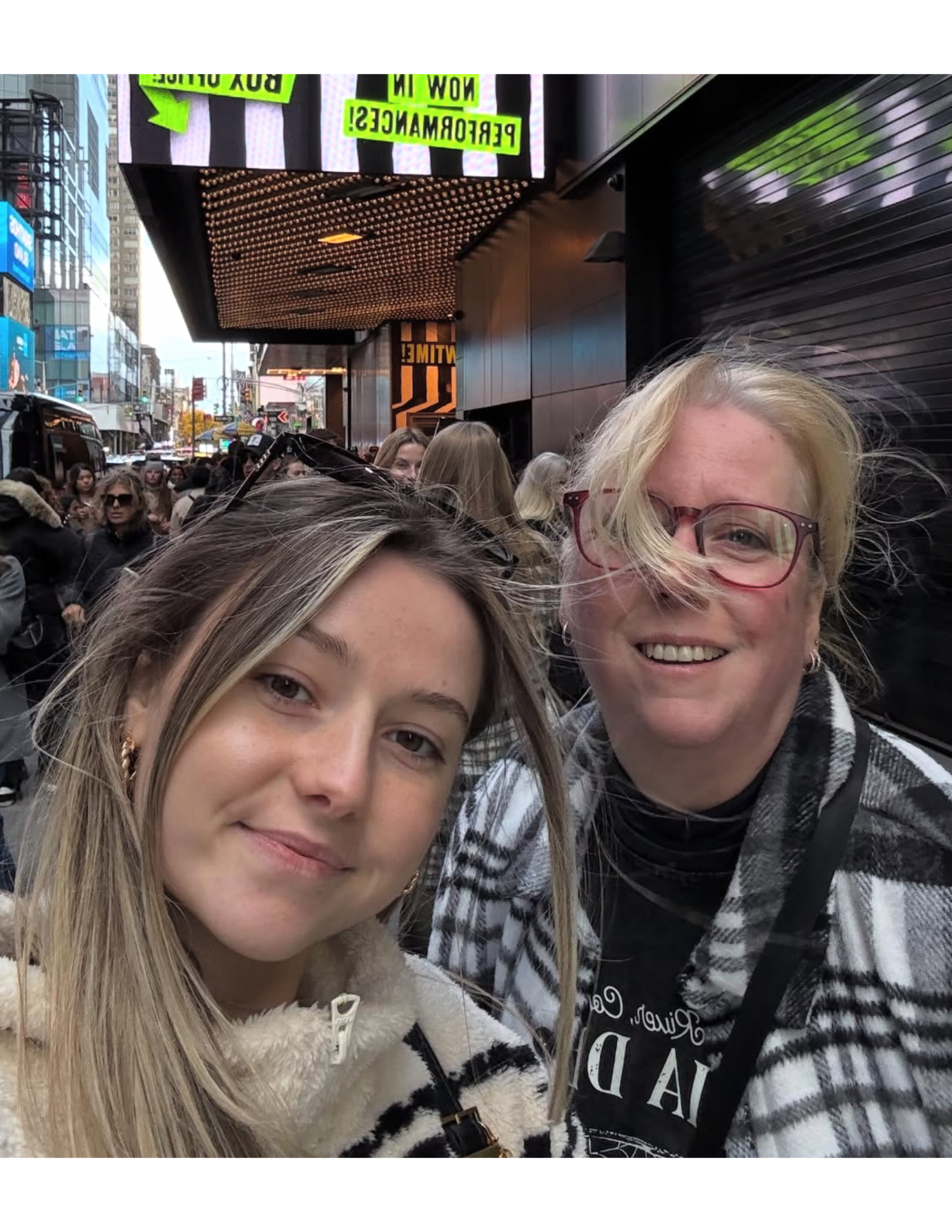 Two women taking a selfie on a busy city street with many people, tall buildings, and digital billboards in the background.