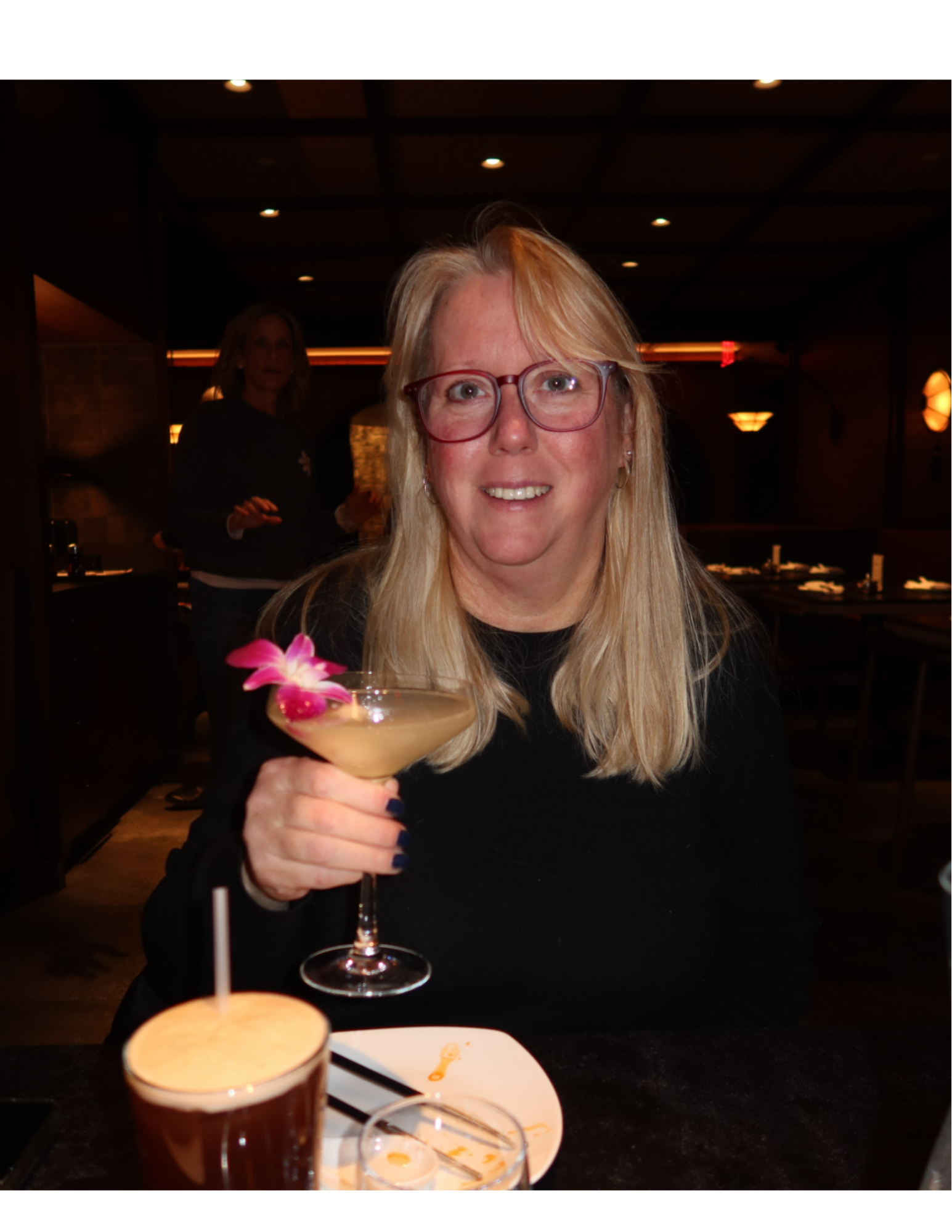 A woman with blonde hair, glasses, and earrings smiling and holding a tropical cocktail with a pink flower garnish in a restaurant or bar setting. In the foreground, there is a dark beer with a frothy head. In the background, another woman is visible, slightly blurred, with a dark interior and warm lighting.