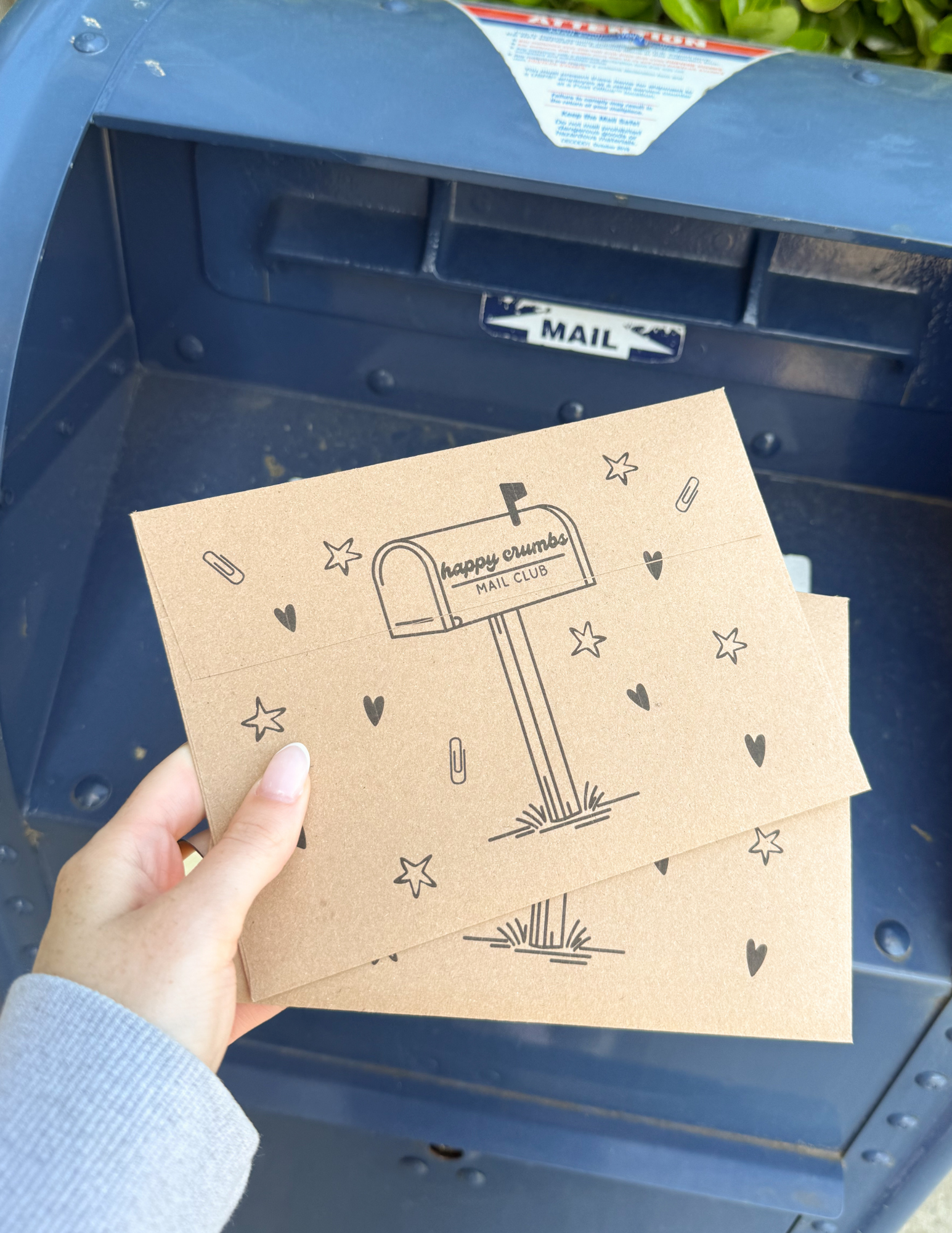 Person holding two brown envelopes with a mailbox illustration and the words "happy crumbs MAIL CLUB" printed on one, inside a blue mailbox.