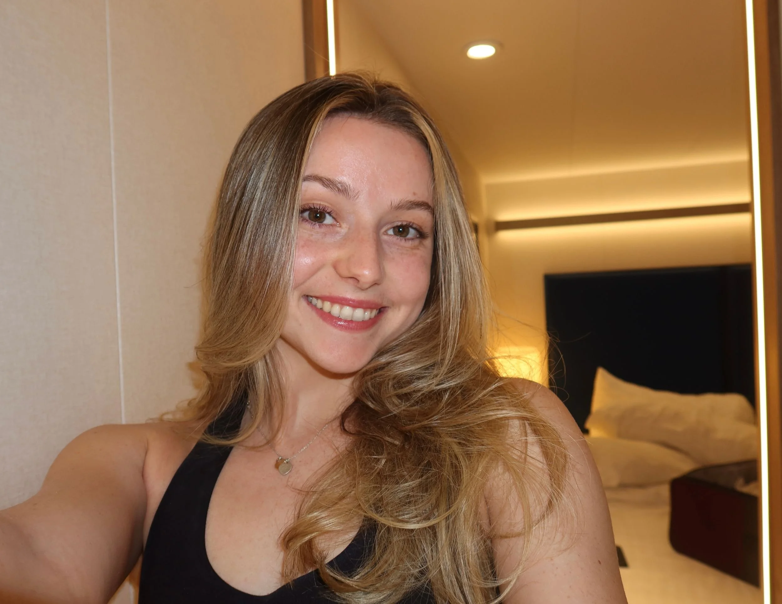 Close-up of a smiling young woman with long, wavy blonde hair, wearing a black top and a silver necklace, indoors with warm lighting and modern decor.