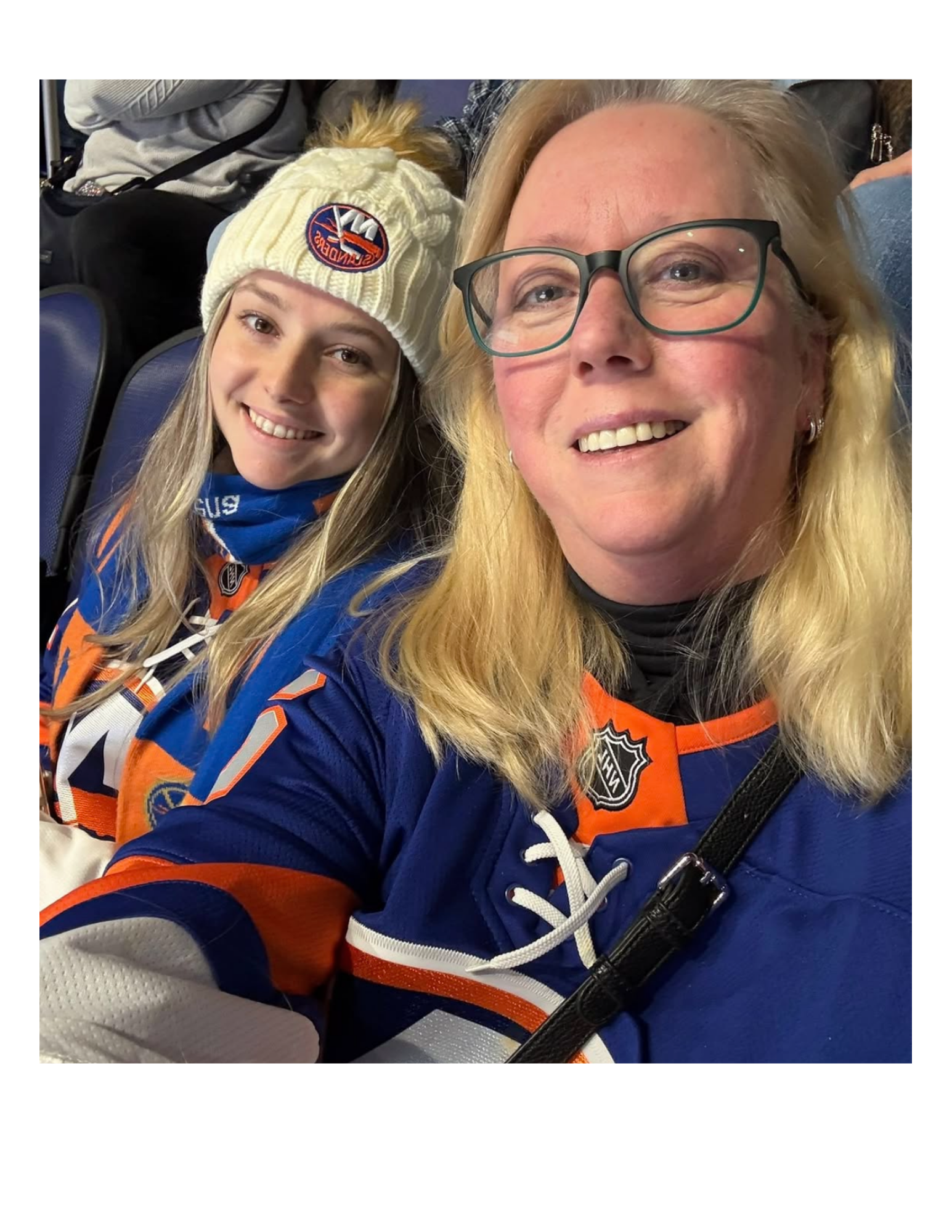 A woman and a young girl taking a selfie at a sports event. The woman is wearing glasses and an NHL hockey jersey, and the girl is wearing a knit hat with a logo and a sports jacket.