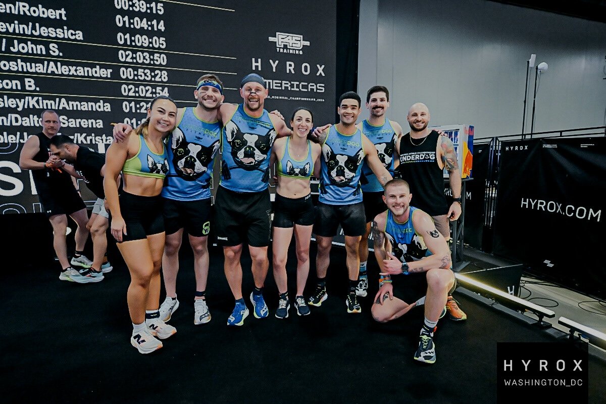 Group of seven Underdog Fitness athletes in colorful matching workout outfits posing together at HYROX, with a large screen background displaying event details and timer, and a few other people in the background.