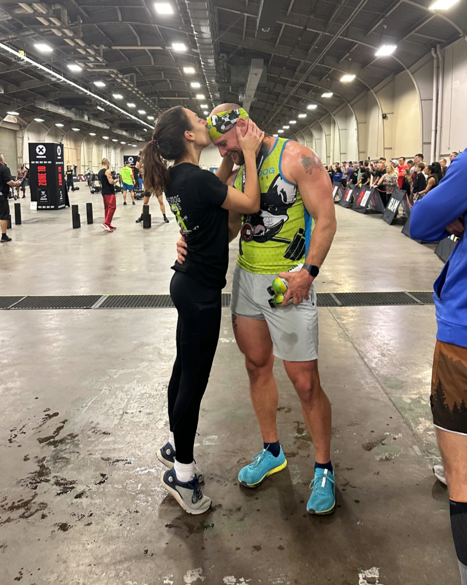 A woman and a man sharing an emotional moment in an indoor sports arena, with the woman holding the man's face and both appearing exhausted and happy after a race or athletic event, with spectators and event setup in the background.