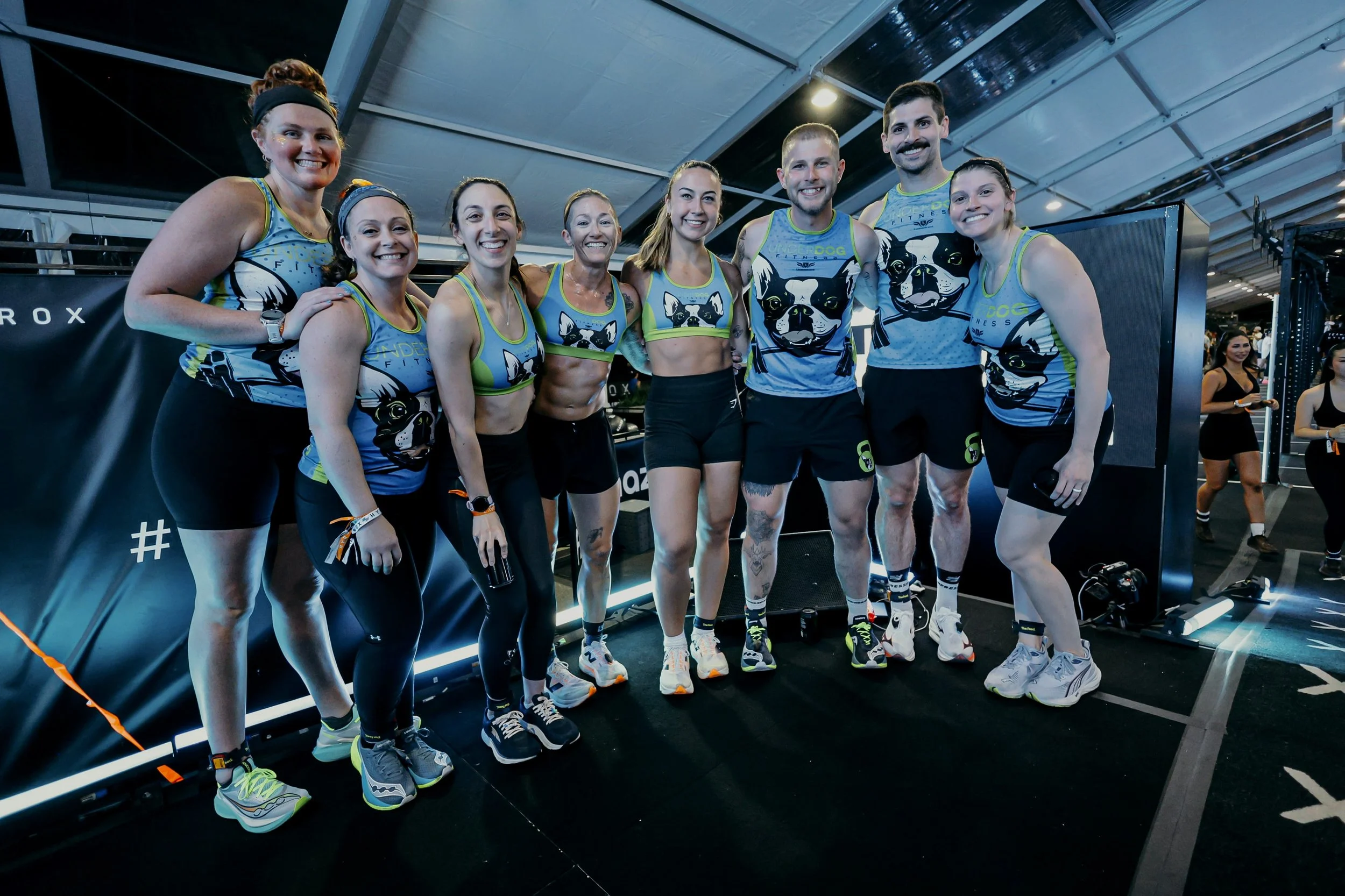 Group of eight smiling people in colorful athletic outfits standing together inside a gym or fitness facility.