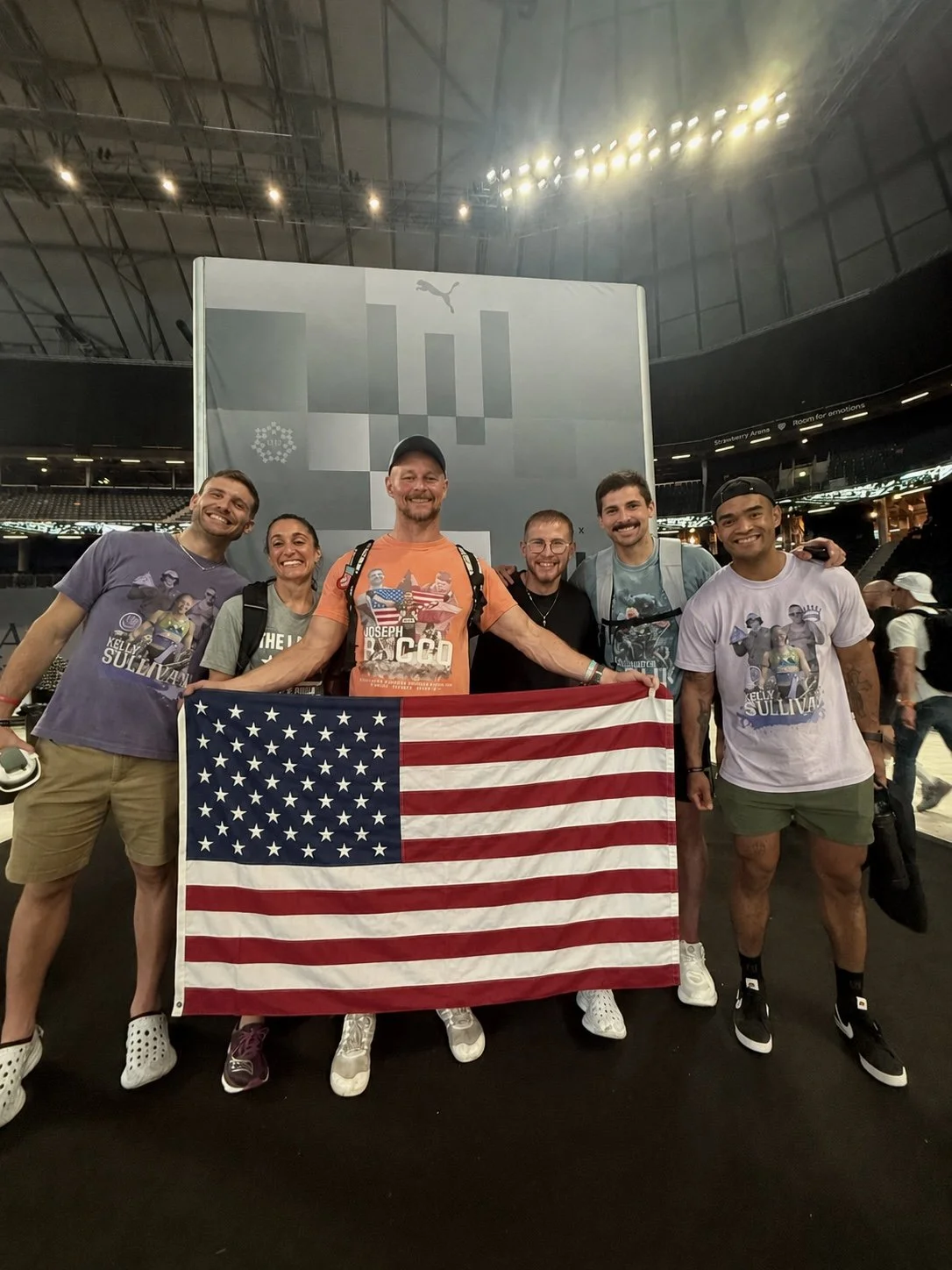 Group of six people smiling and holding an American flag inside a large indoor stadium, with a large screen in the background displaying Puma logo. Photo taken at the 2026 HYROX World Championship.