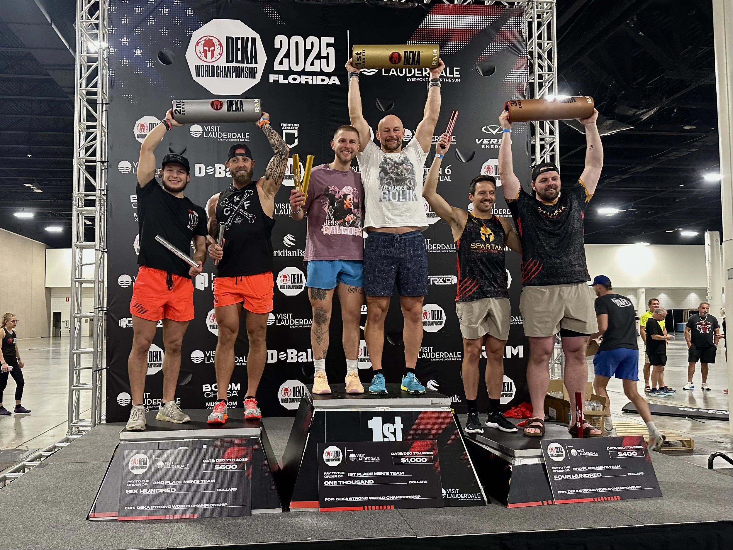 Six men standing on an award podium with medals and trophies, celebrating their victory at the DEKA World Championship 2025 in Florida; one in the center holding a large gold-colored trophy, with a backdrop displaying event information.