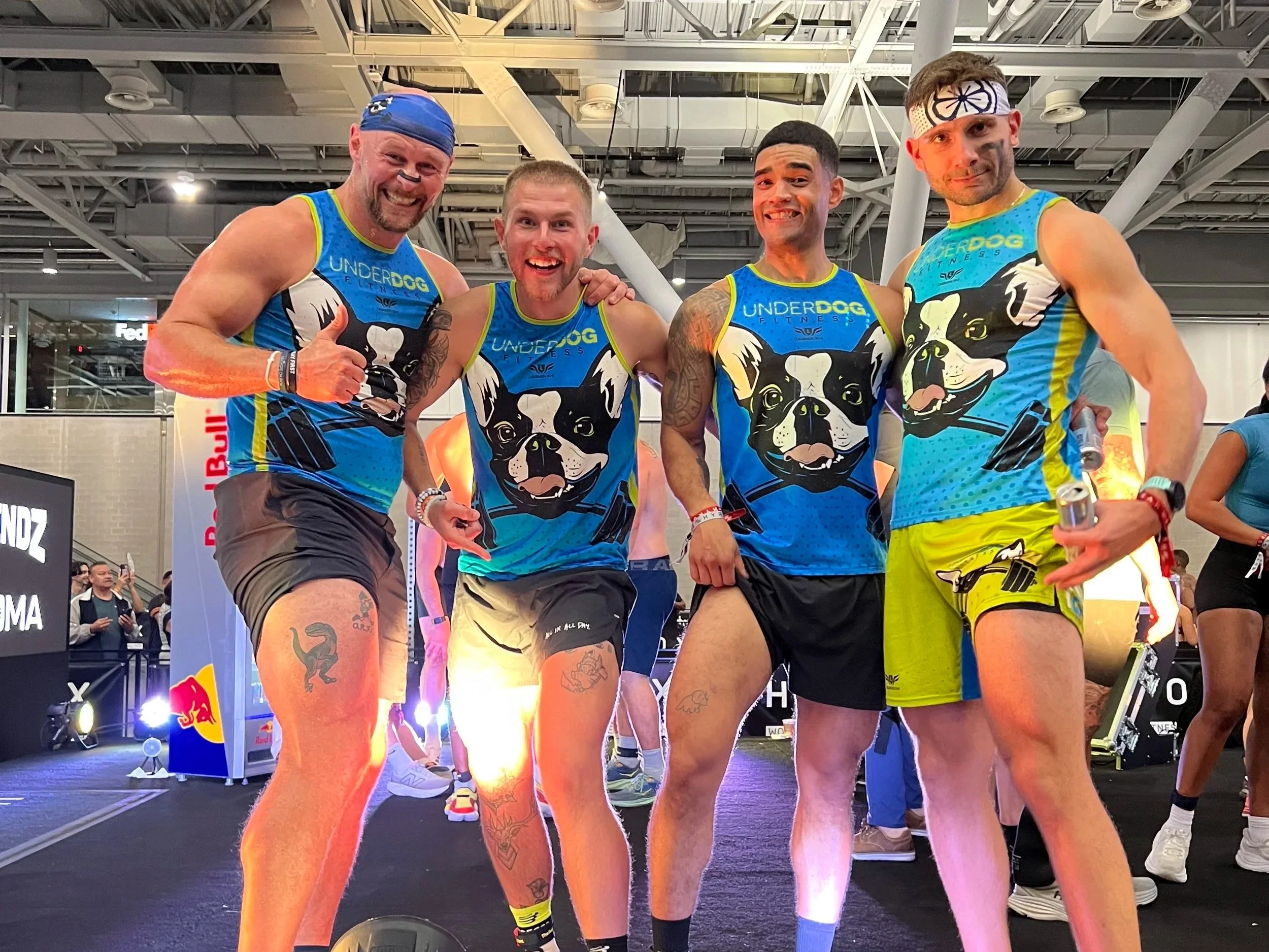 Four muscular men in matching blue tank tops with a dog logo, posing and smiling at an indoor fitness event. They are wearing athletic shorts and wristbands, with one giving a thumbs-up.