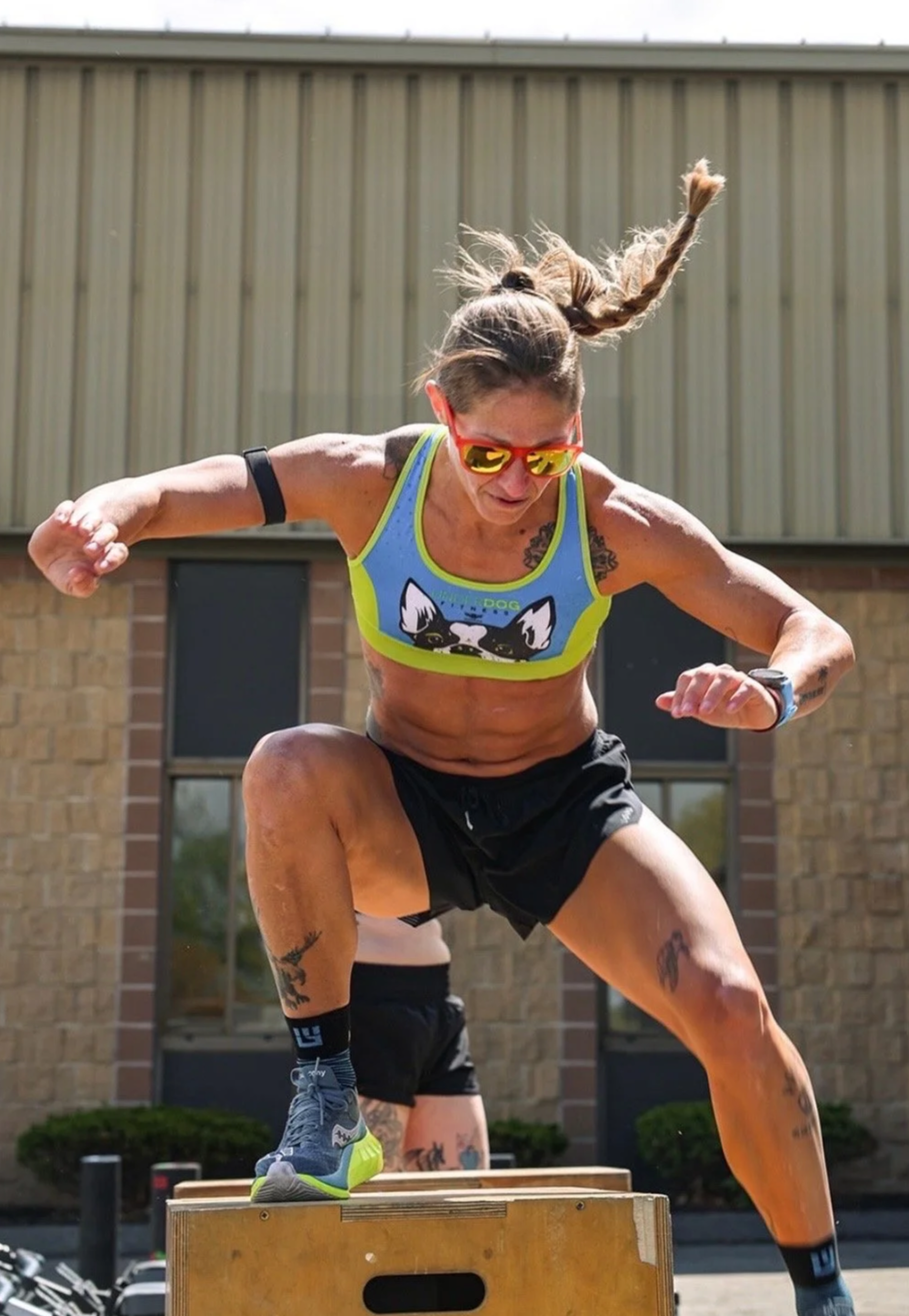 A woman in athletic gear jumps onto a wooden box during an outdoor workout, wearing sunglasses and a tank top with a dog graphic.