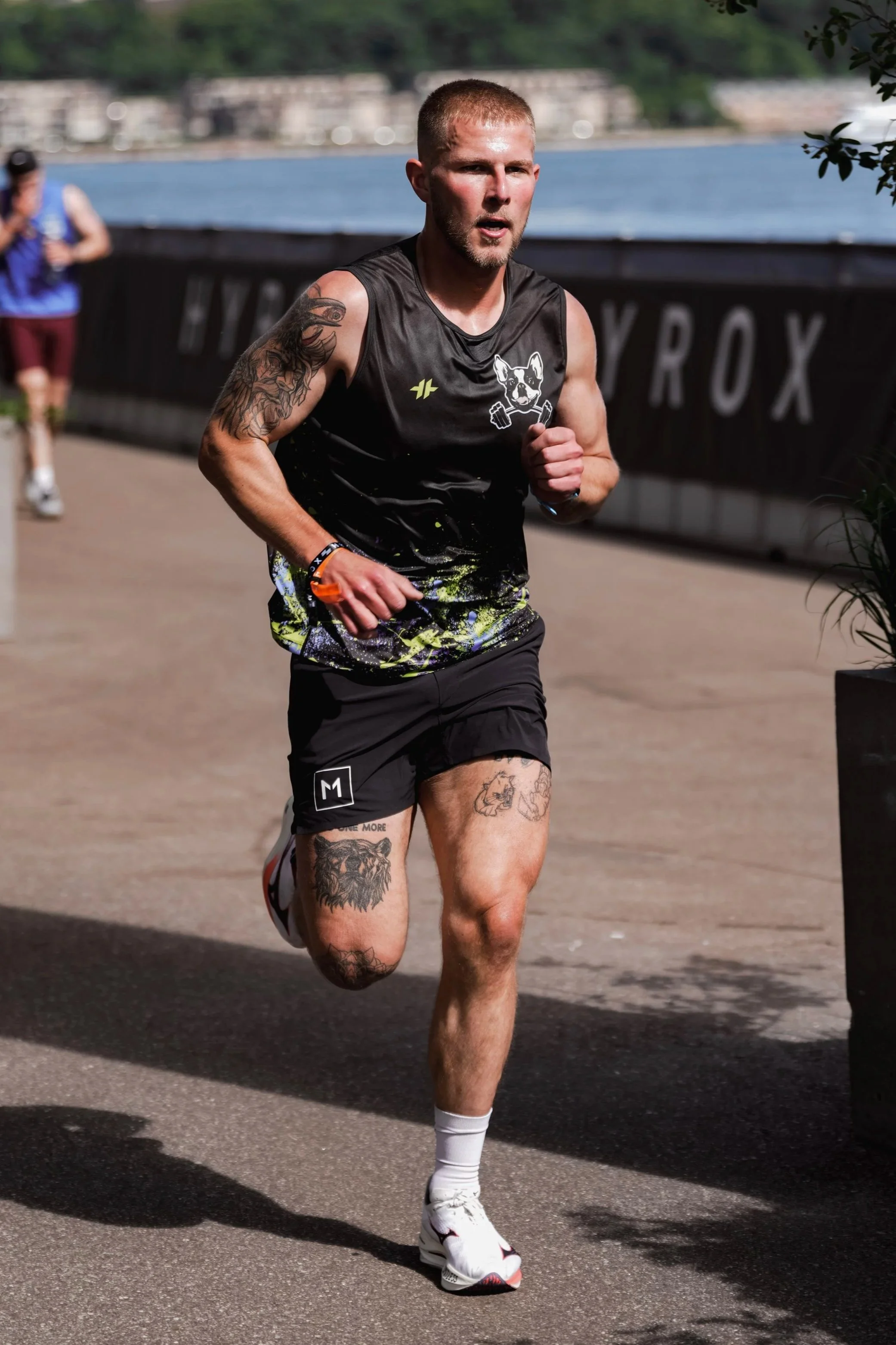 A man running outdoors along a paved path near a body of water, wearing a black sleeveless athletic shirt, shorts, and white running shoes, with tattoos on his arms and legs.