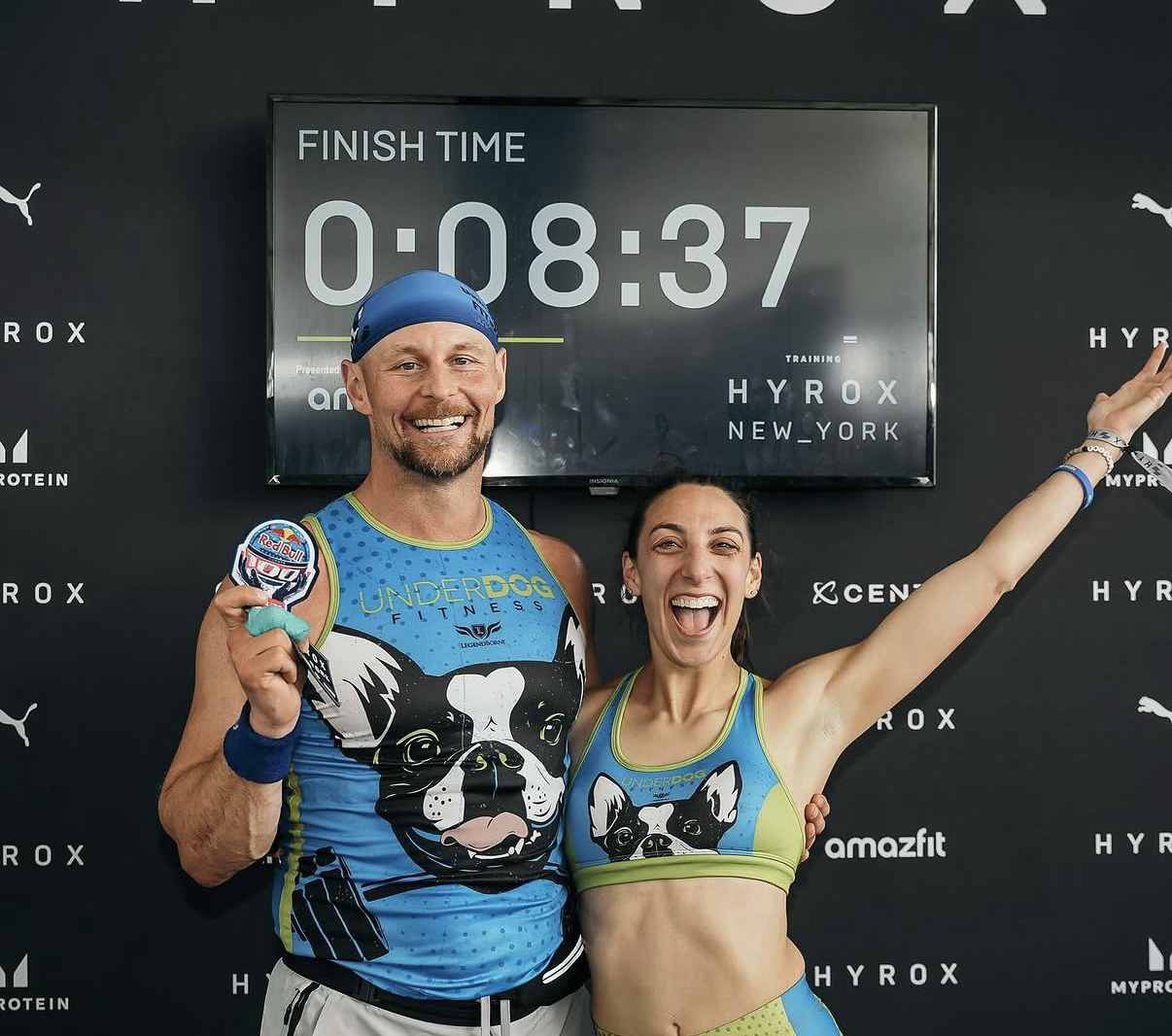 Two HYROX runners, a man and a woman, smiling and celebrating after a race with a finish time. They are wearing matching Underdog Fitness athletic attire with a French bulldog design and holding medals.