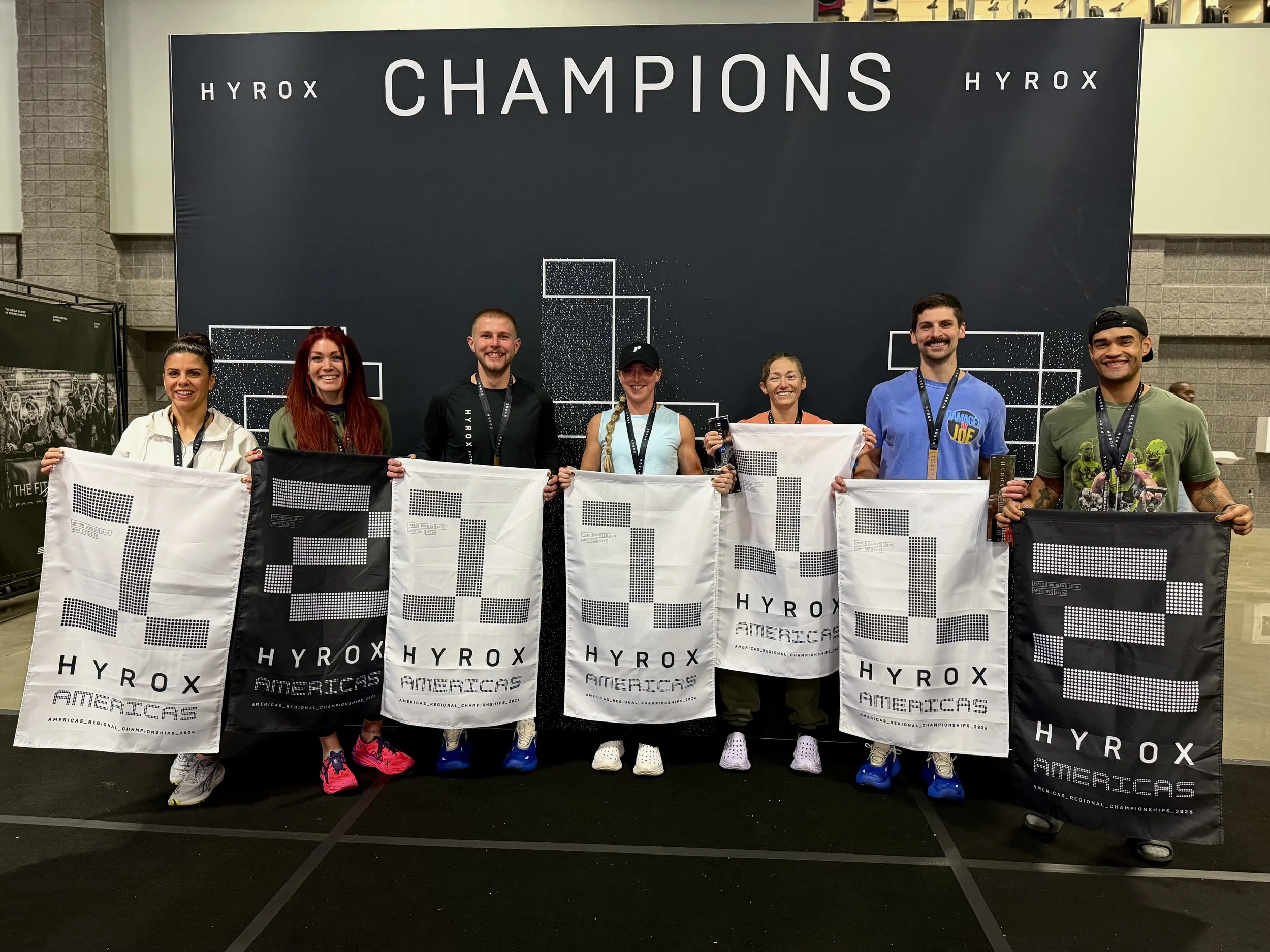 Group of six people standing in front of a backdrop that says 'HYROX Champions,' holding flags with the HYROX logo and text reading 'Americas'. They are wearing athletic clothing and medals, smiling for the photo.