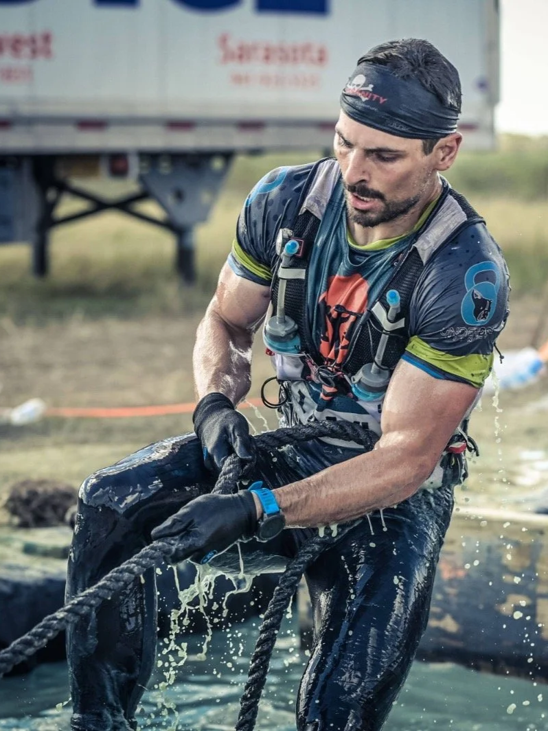 Male athlete in black gloves pulling on a rope, wearing a headband, wet athletic clothing, and hydration gear, participating in an outdoor obstacle course race.