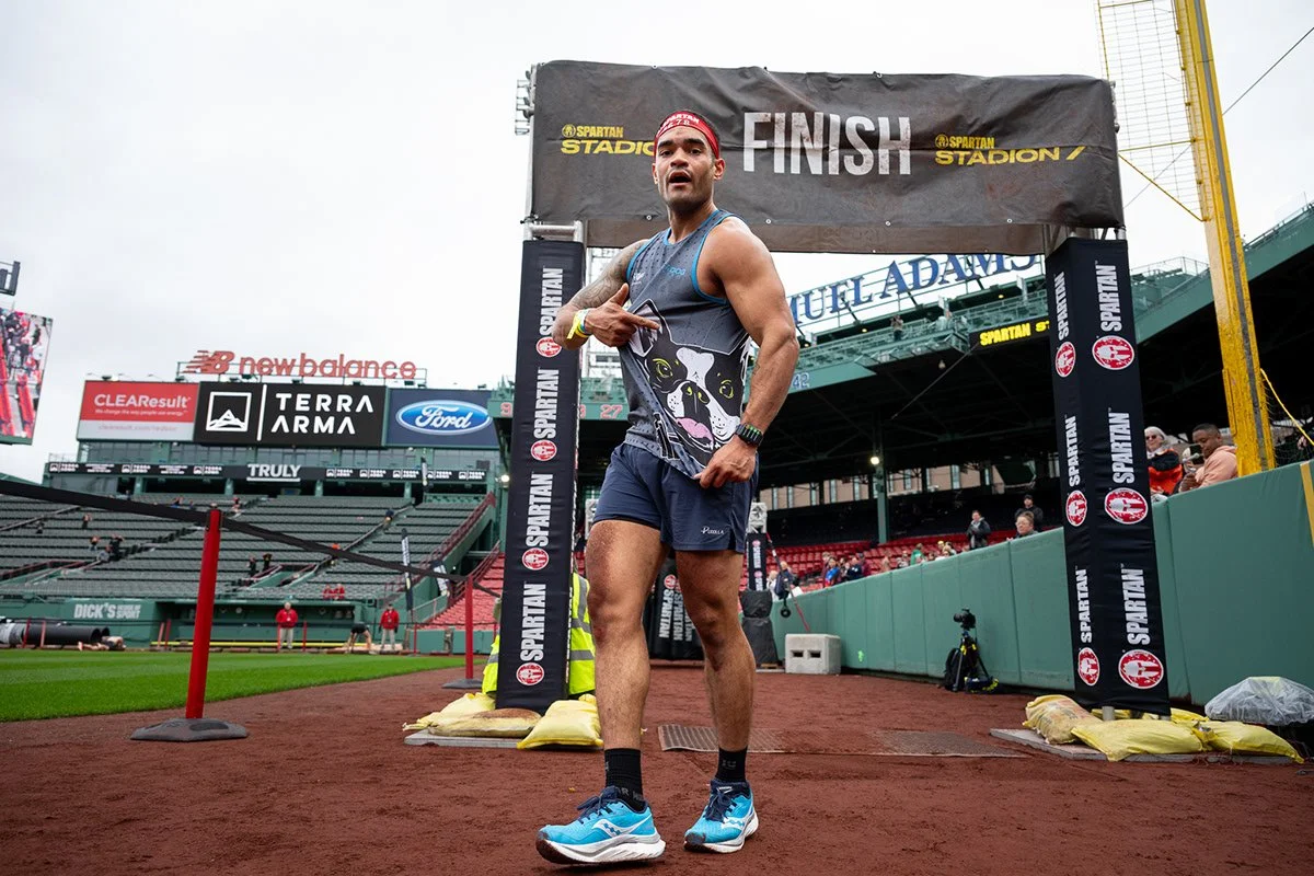 A male marathon finisher standing at the race finish line, pointing at himself, wearing a tank top, shorts, and running shoes, with a finish banner overhead at a stadium.