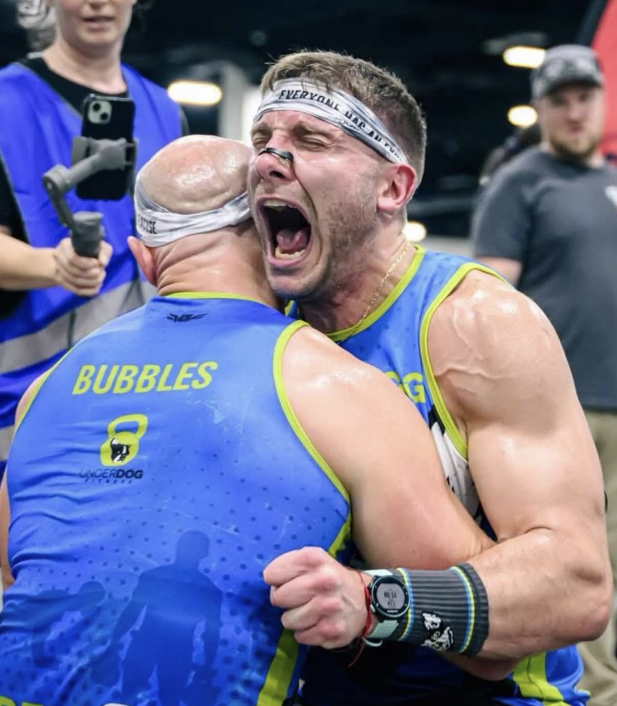 Two male athletes in blue sports jerseys with yellow trim celebrating passionately after a sports event, embracing each other tightly, with one appearing to shout or yell in excitement.