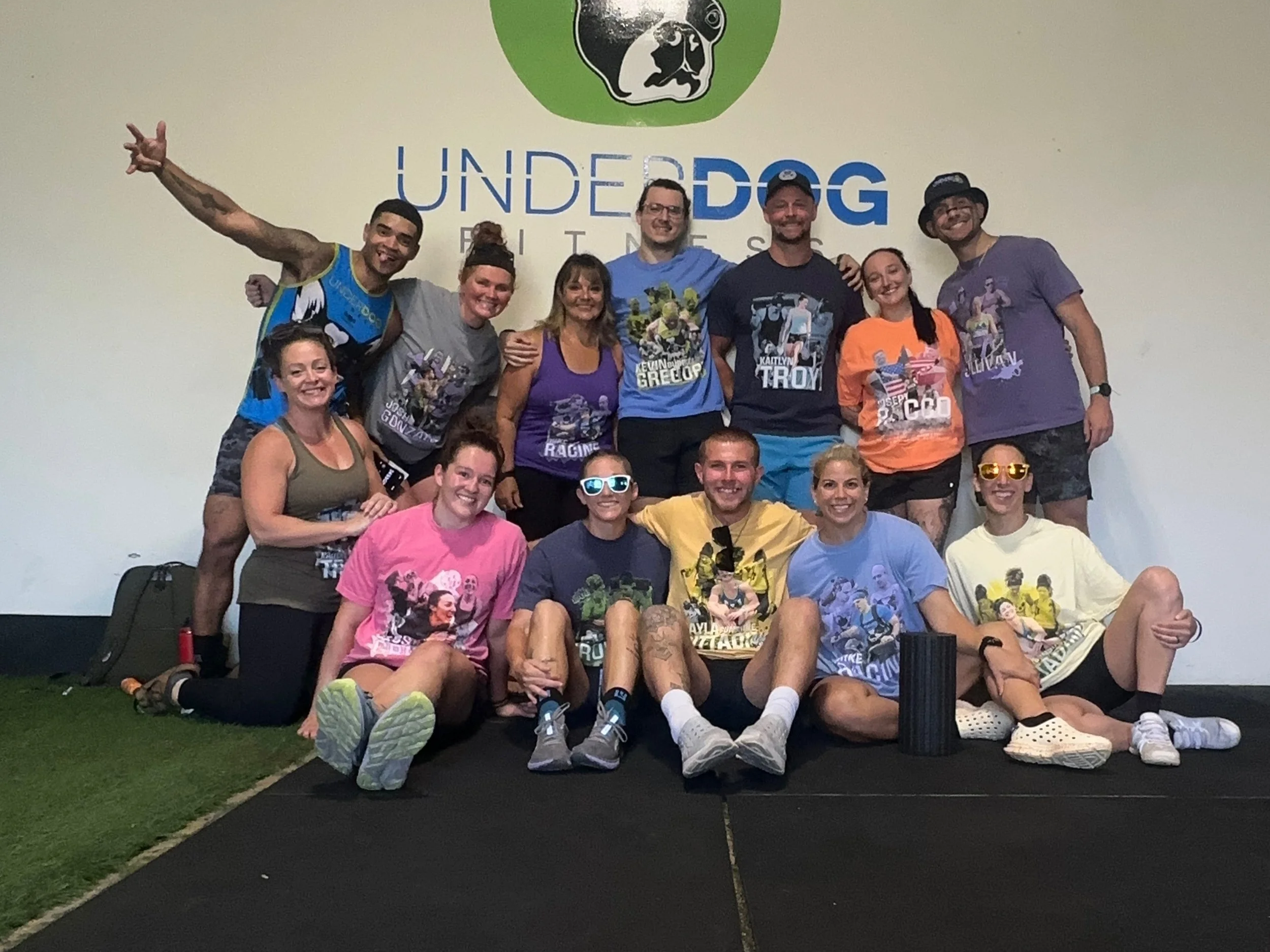 Group of people posing for a photo indoors in front of a sign that reads 'Underdog RACING' with a graphic of a dog's face. The group includes men and women, some standing and some sitting or kneeling, mostly smiling and wearing casual athletic clothing.
