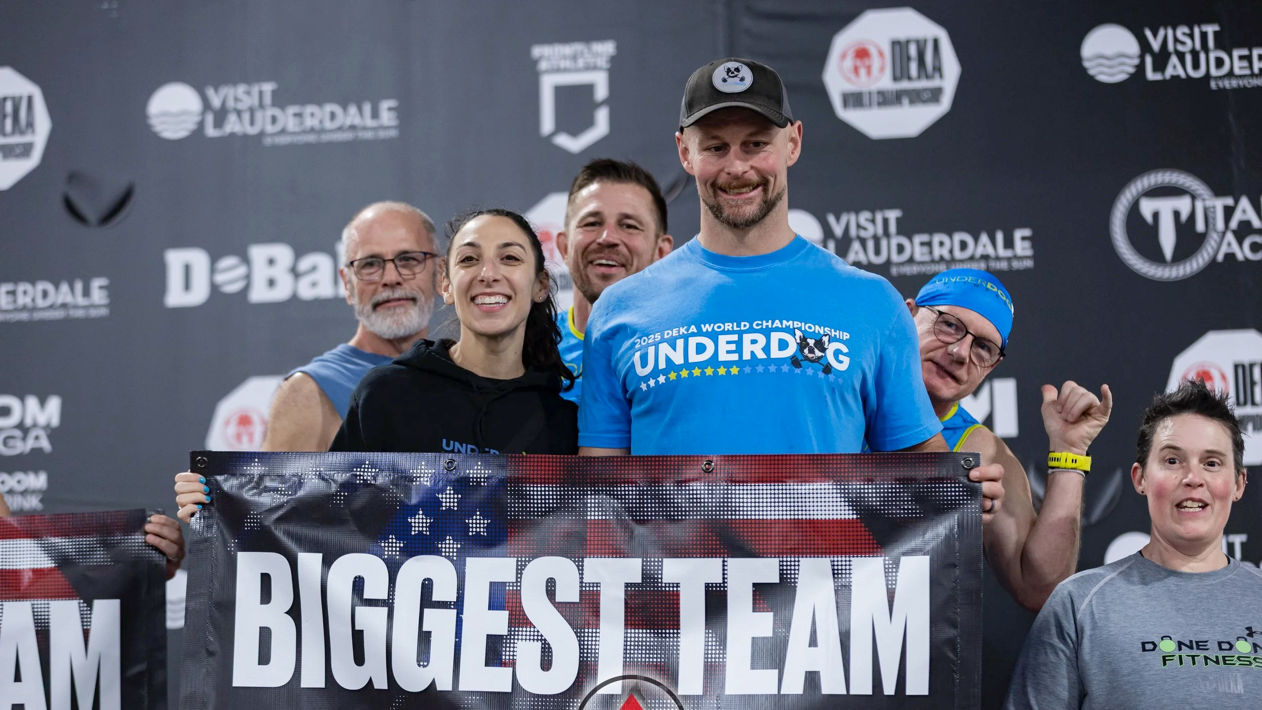 Group of people at a competition, with the man in the center wearing a blue shirt that says '2025 Deka World Championship UNDERDOG' and a black baseball cap, smiling and posing for the photo, with others around him also smiling and holding a banner.