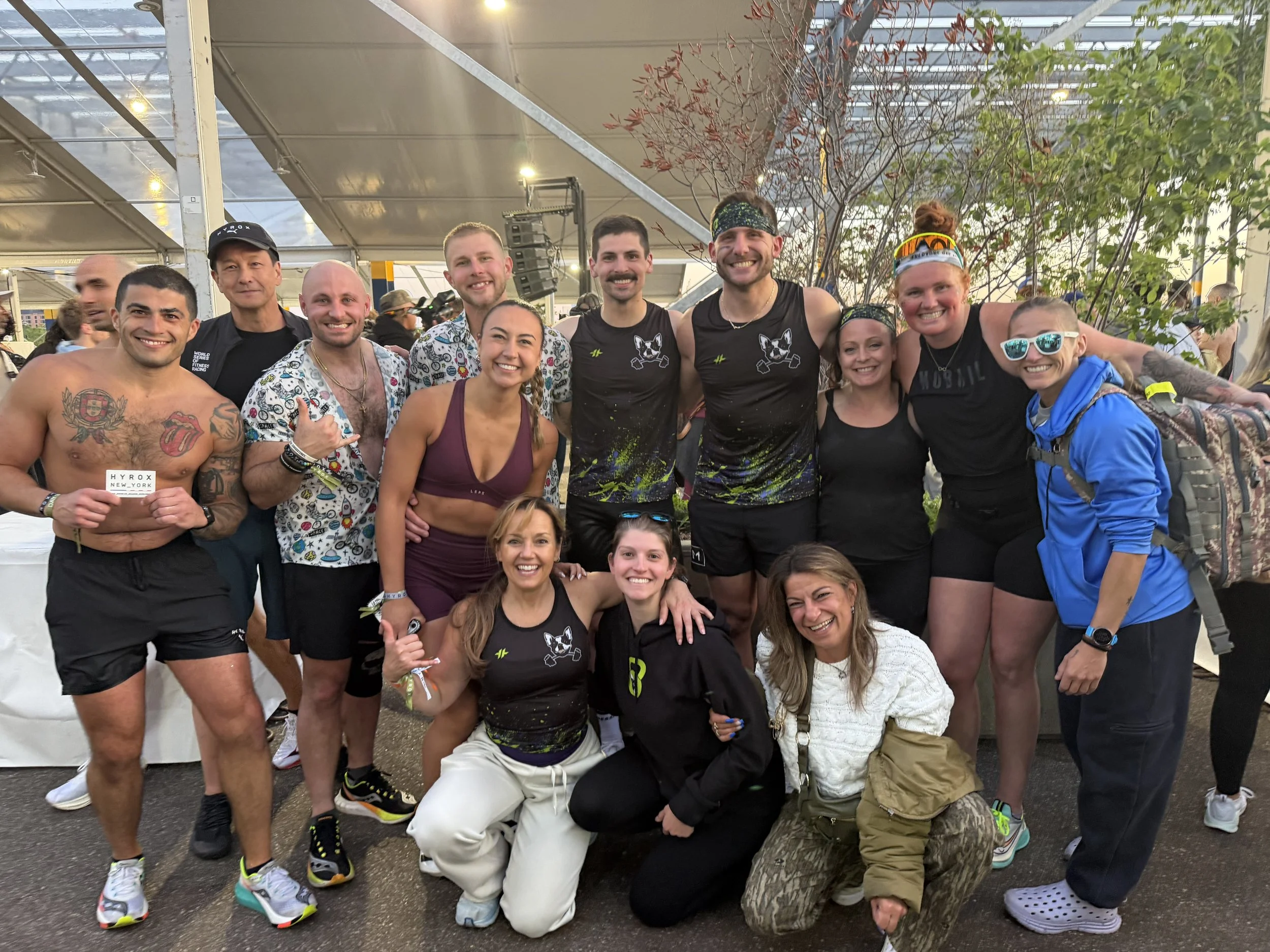 A large group of smiling people in athletic attire at a race event, standing together inside a large tent with metal structure and trees outside.