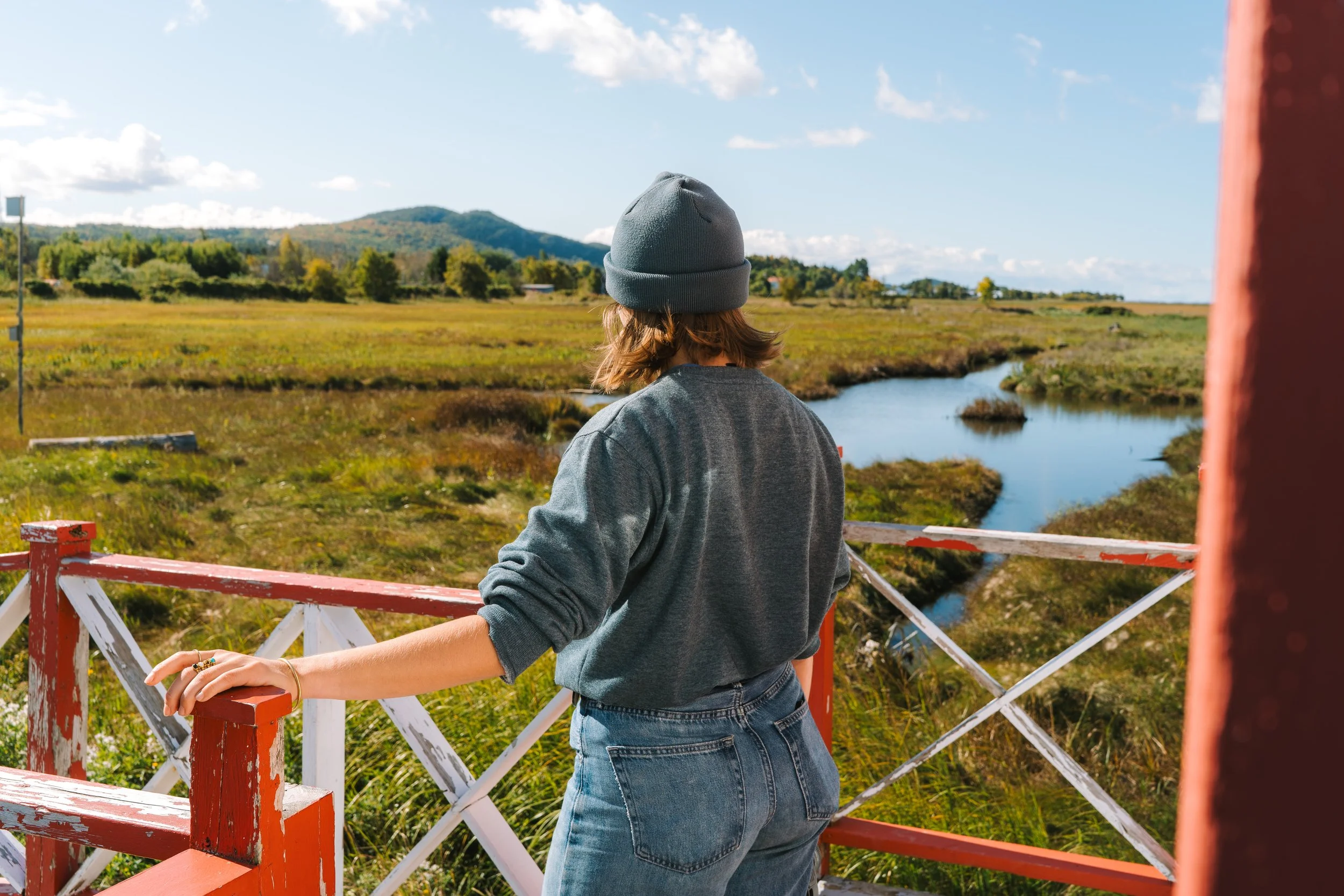A girl wearing a blue hoodie and beanie hat, standing on a red and white wooden bridge in Kamouraska overlooking a grassy marshland with a winding creek, trees, and hills in the distance under a blue sky with scattered clouds.