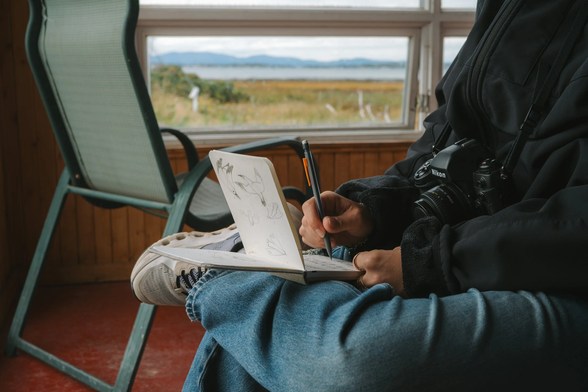 Person sitting indoors near a window with a view of mountains and water, drawing or writing in a notebook with a pen, wearing a black jacket and blue jeans.