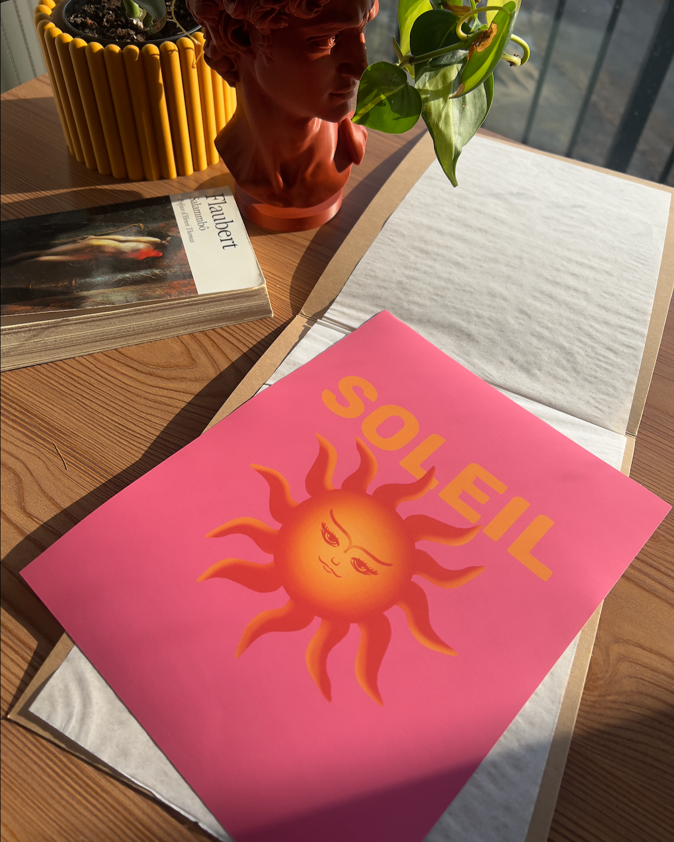 A wooden table with a dopamine pink poster featuring a stylized sun with a woman's face, a small potted plant in a reddish-brown bust-shaped planter, a book titled "Haubert" on the table, and some sheets of paper in the sunlight.